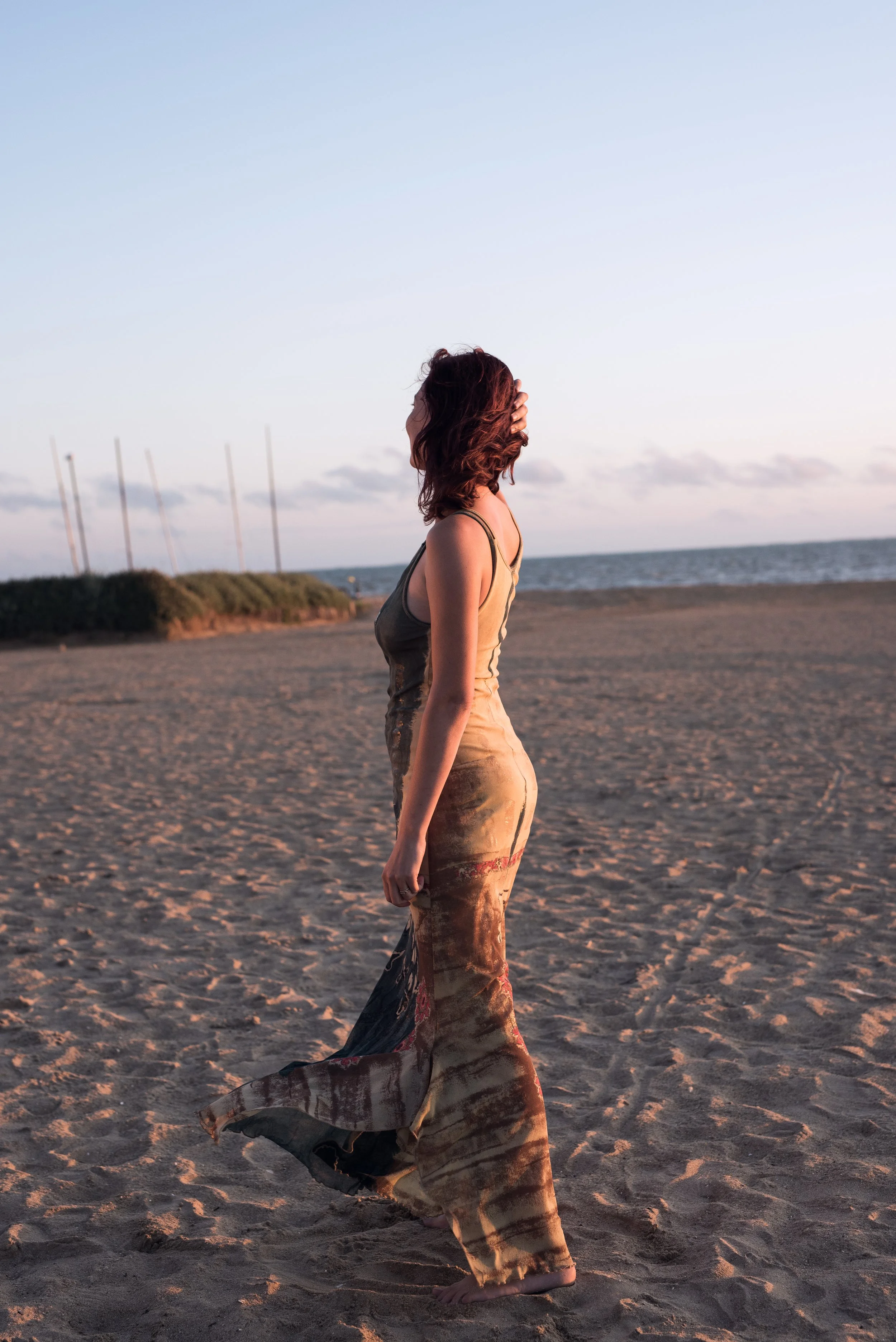 Femme avec cheveux roux, portant une robe de couleur beige et un manteau long à motif floral, marche pieds nus sur la plage au coucher du soleil.