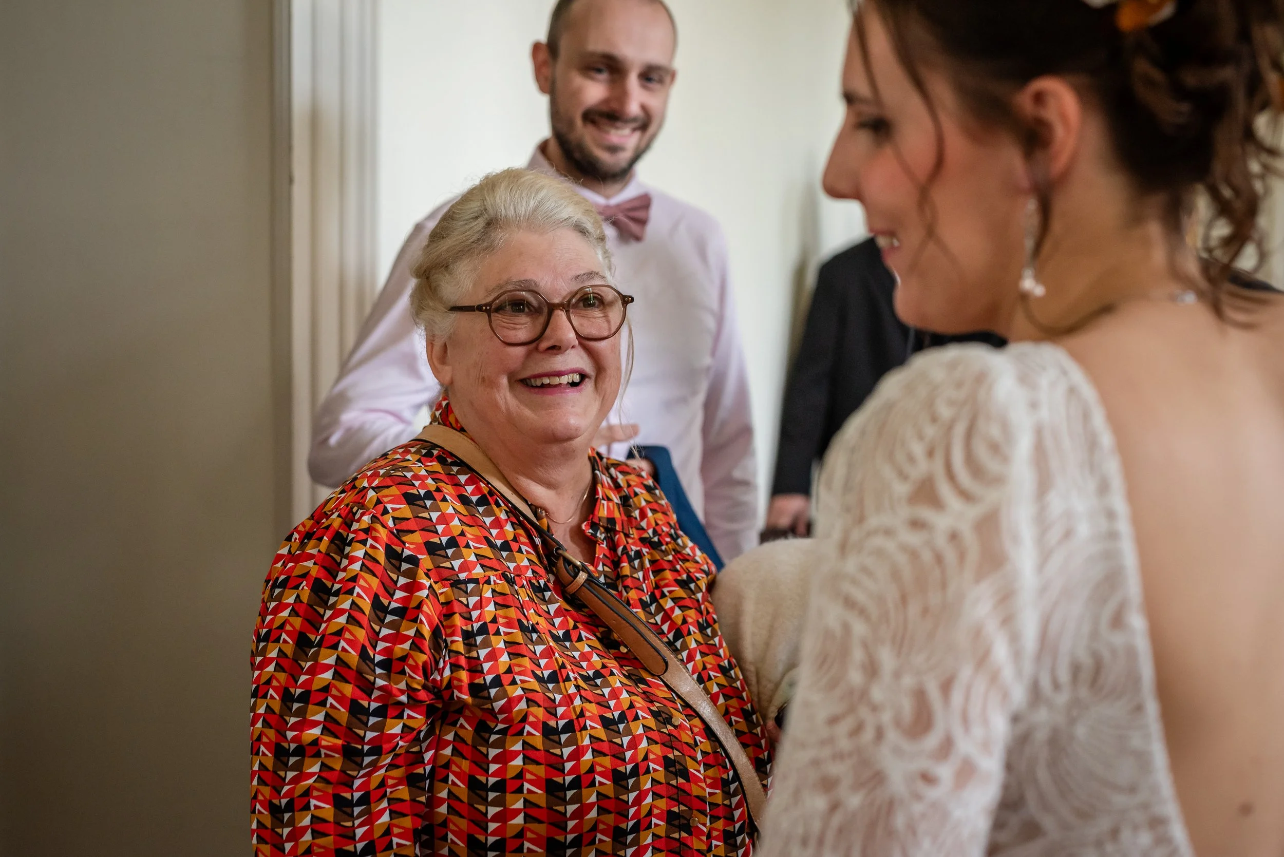 Une femme âgée portant des lunettes sourit, parlant à une jeune femme dont la robe en dentelle est visible. Deux hommes derrière, l'un portant une chemise à col et une cravate rose, sourient et regardent la scène.