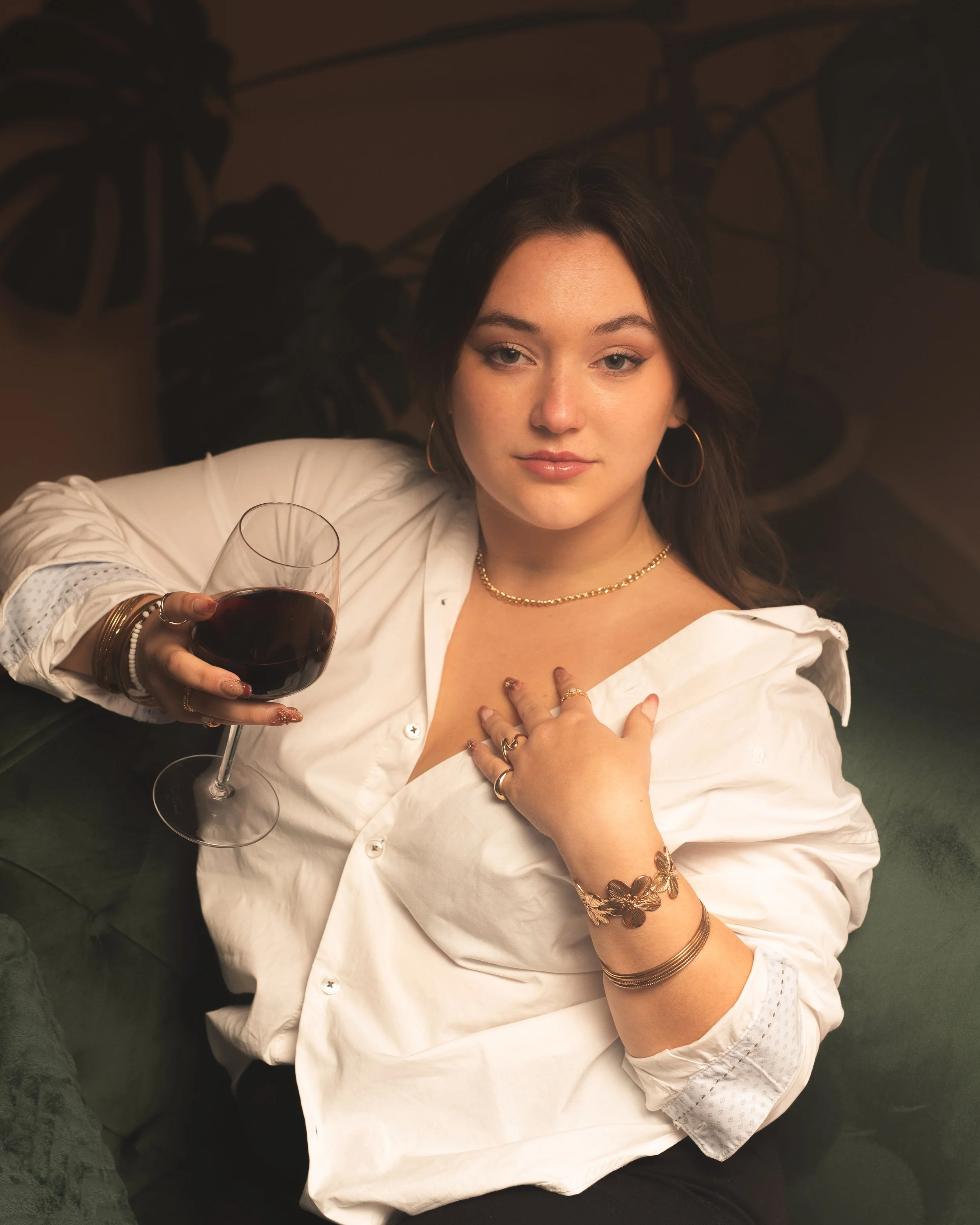 Une femme avec des cheveux bruns longs et bouclés, portant une chemise blanche avec des détails en dentelle, portant plusieurs bracelets et une chaîne en or, tenant un verre de vin rouge, assise dans un environnement intérieur sombre avec des plantes