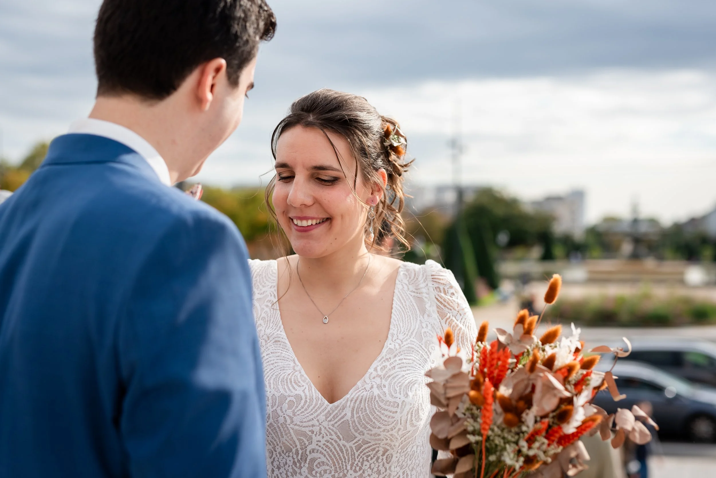 Femme en robe blanche tenant un bouquet de fleurs, échangeant un regard avec un homme en costume bleu, lors d'une occasion spéciale en plein air.
