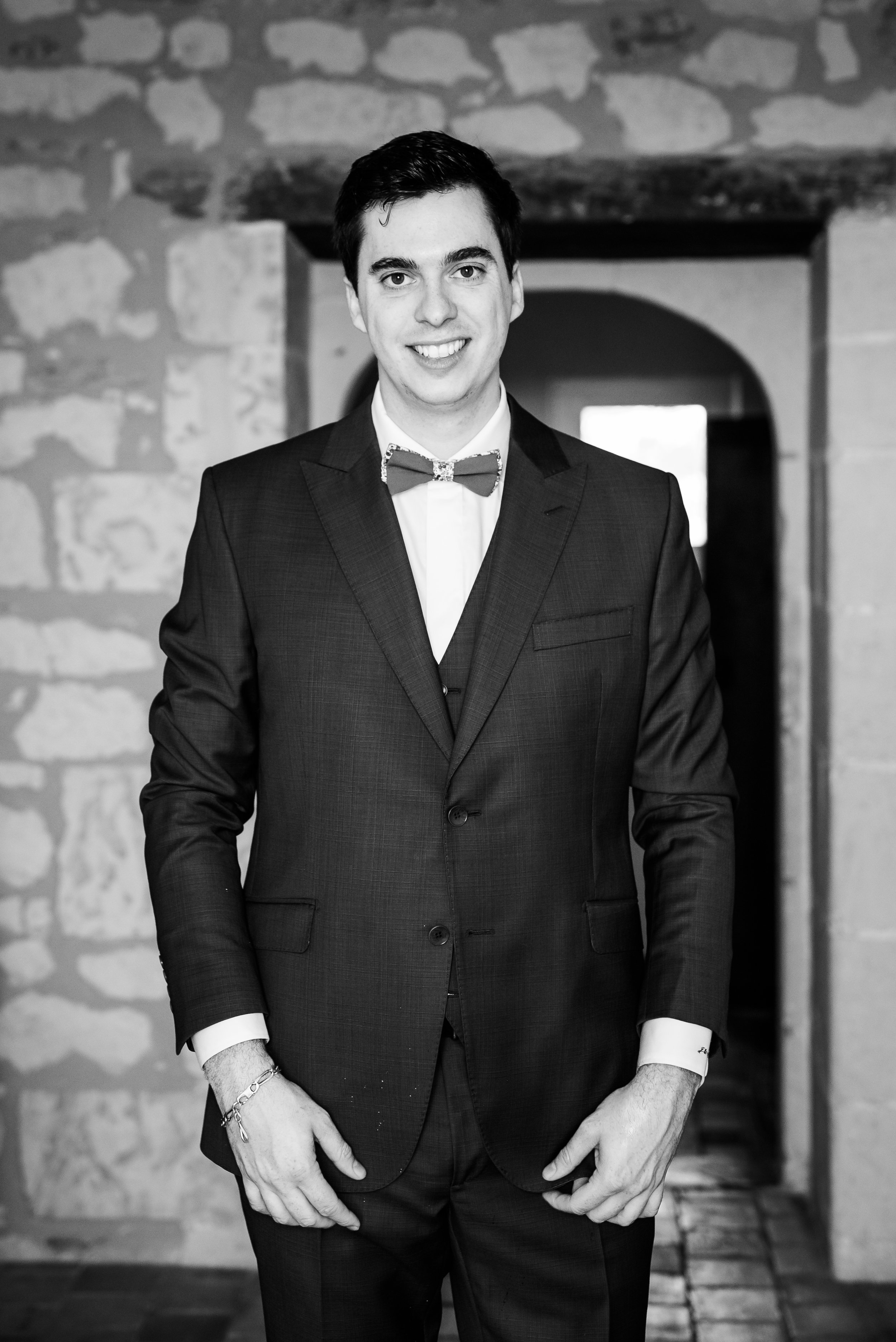 Un jeune homme en costume noir et nœud papillon, sourire, dans un lieu avec mur en pierre, en noir et blanc.