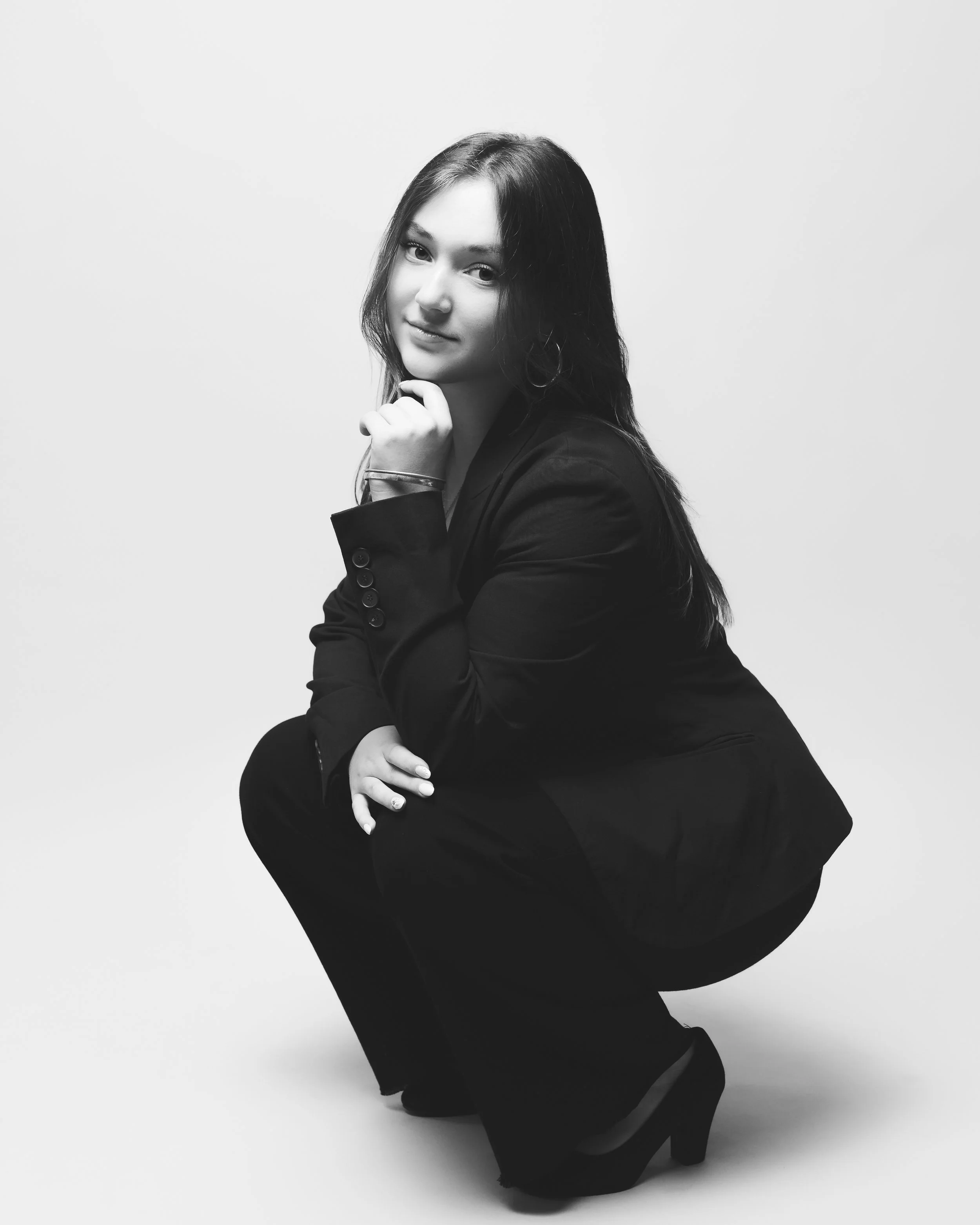 Une femme en noir et blanc, à genoux, portant un blazer noir et des talons hauts, regardant vers l'appareil et posant sa main sous son menton.