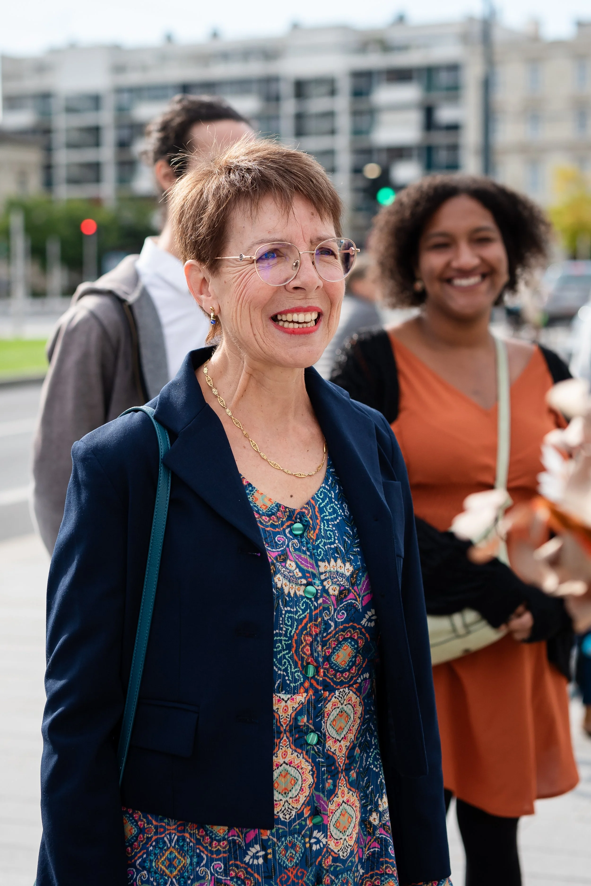 Trois femmes souriantes devant un bâtiment résidentiel, vêtues de vêtements colorés, en plein air, avec une ambiance joyeuse.
