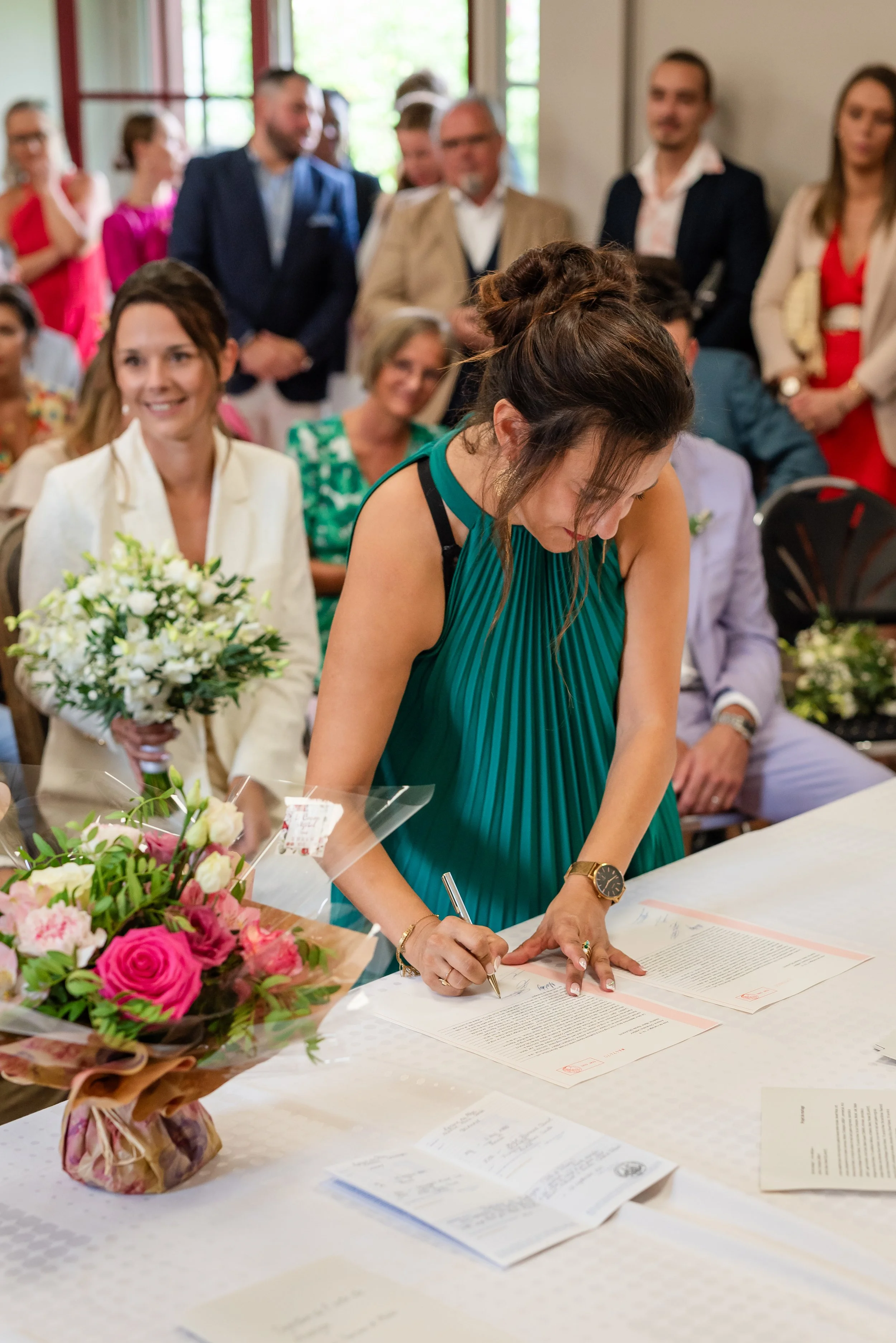 Une femme signe un document lors de ce qui semble être une cérémonie, entourée de plusieurs personnes debout et assises, avec des bouquets de fleurs sur la table.