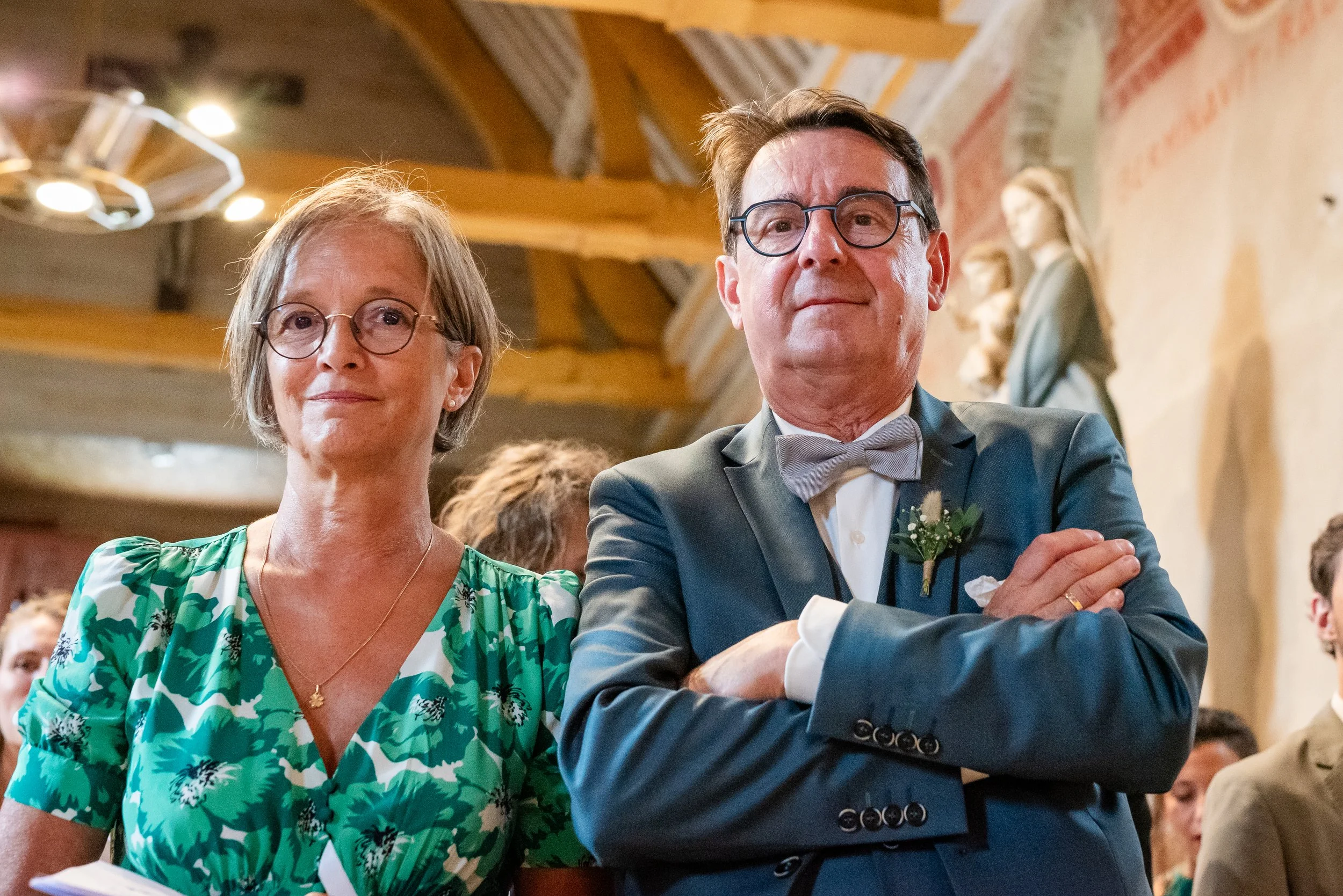 Un homme et une femme assis lors d'un événement, lui en costume avec nœud papillon, elle en robe verte à motifs floraux, tous deux portant des lunettes, dans une salle avec statues et éclairage chaud.