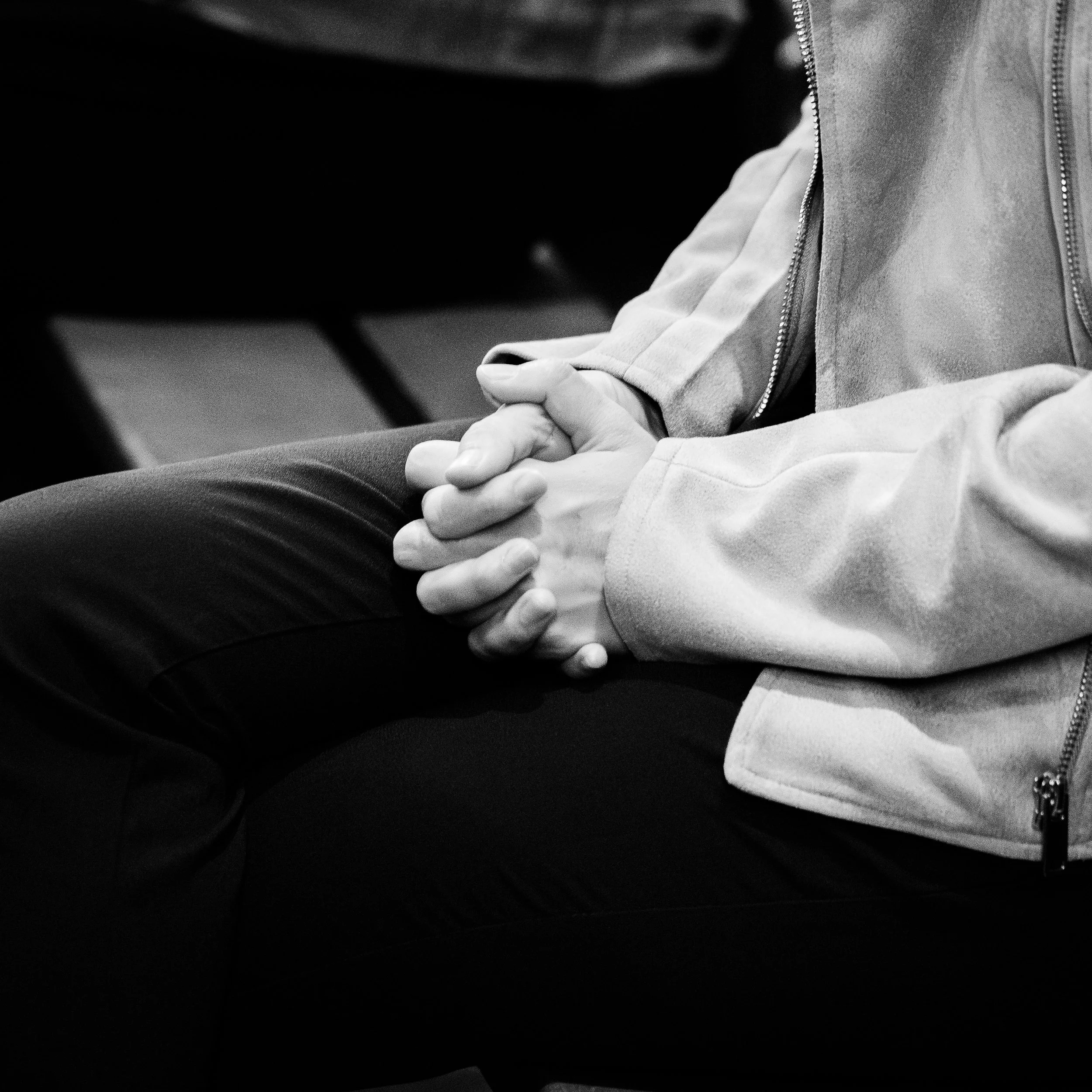Une personne portant une veste en tissu clair et un pull blanc, assise avec les mains jointes. L'image est en noir et blanc, montrant une scène intérieure.