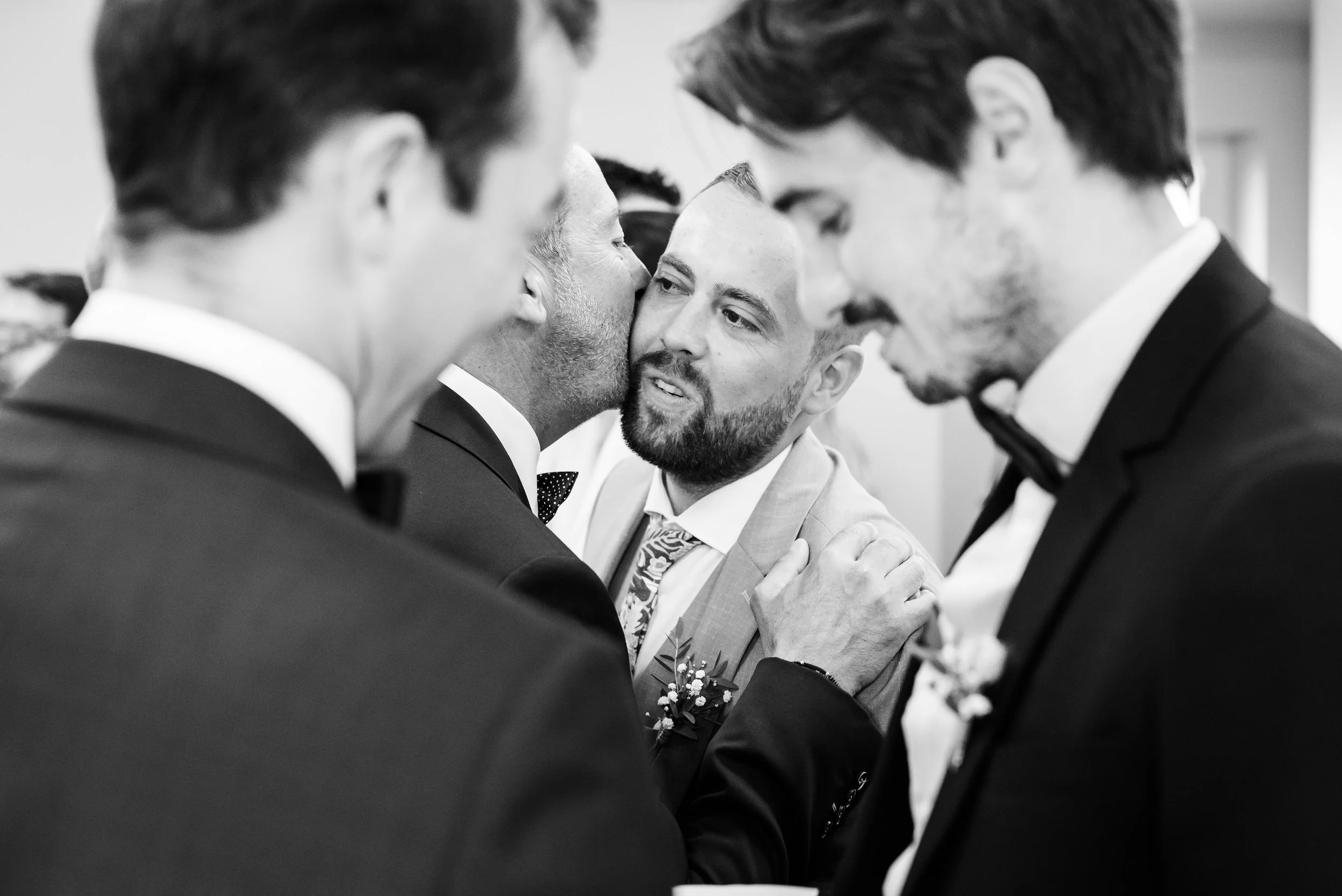 Homme portant un costume et une cheville en bouton roulant ses mains avec un groupe d'hommes en costume tandis qu'il parle à un autre homme, dans un cadre formel.
