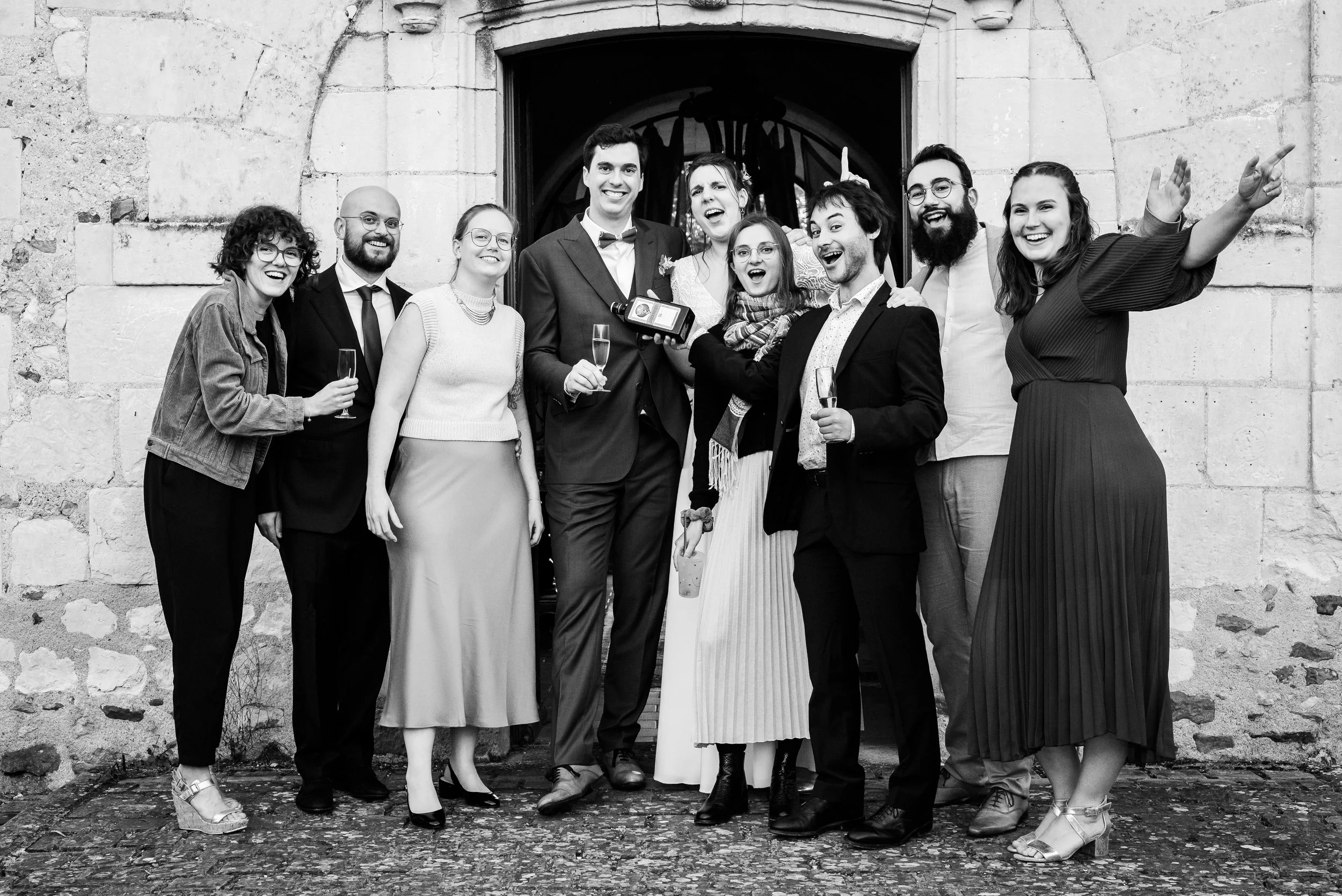 Groupe de personnes souriantes en tenue habillée, posant à l'extérieur devant un bâtiment en pierre, certains tenant des flûtes de champagne, dans une ambiance festive ou célébratoire.