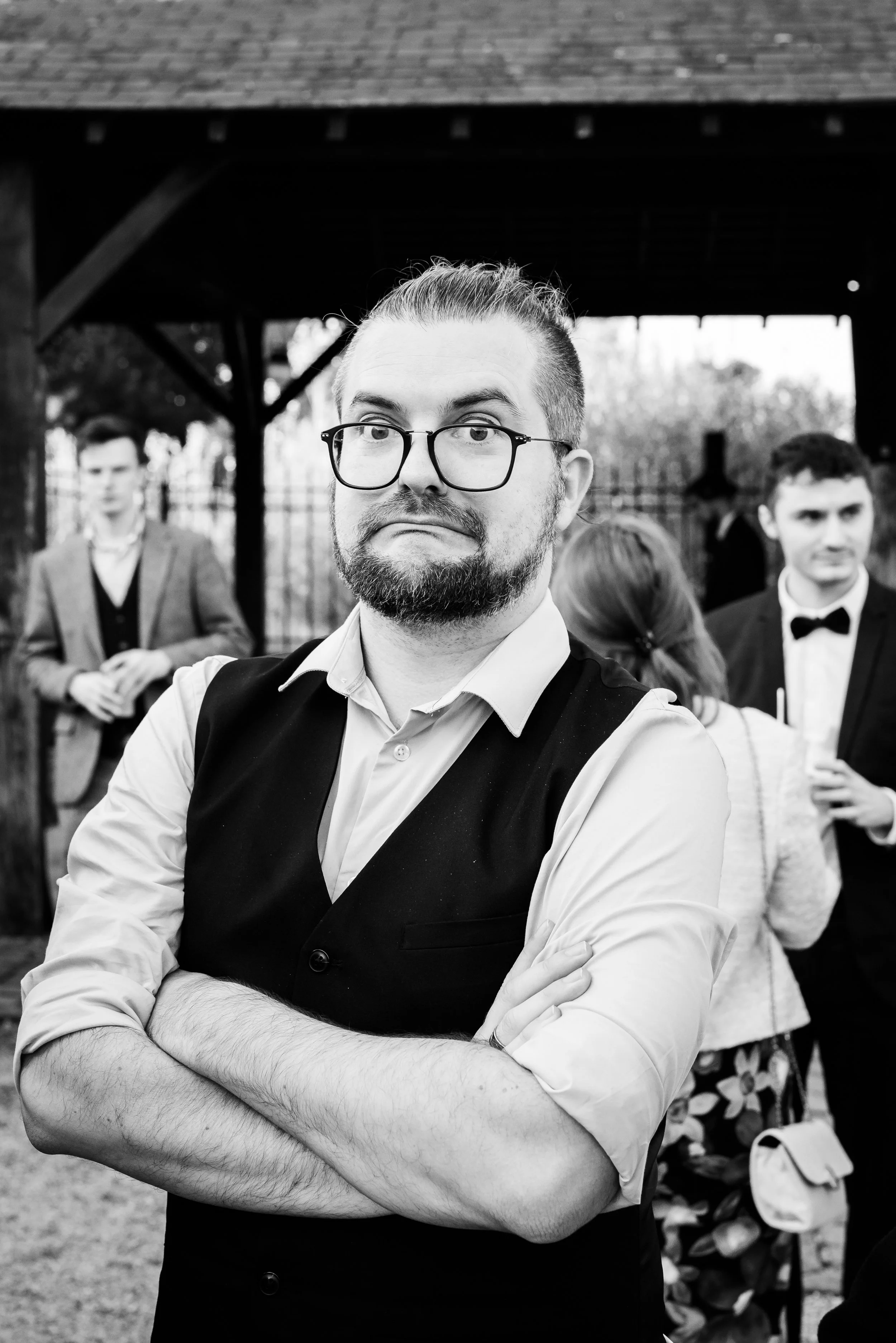 Un homme portant des lunettes, en salopette, avec les bras croisés, en extérieur, lors d'une occasion formelle ou sociale. Autres personnes en arrière-plan, en tenue élégante, sous un abri en bois.