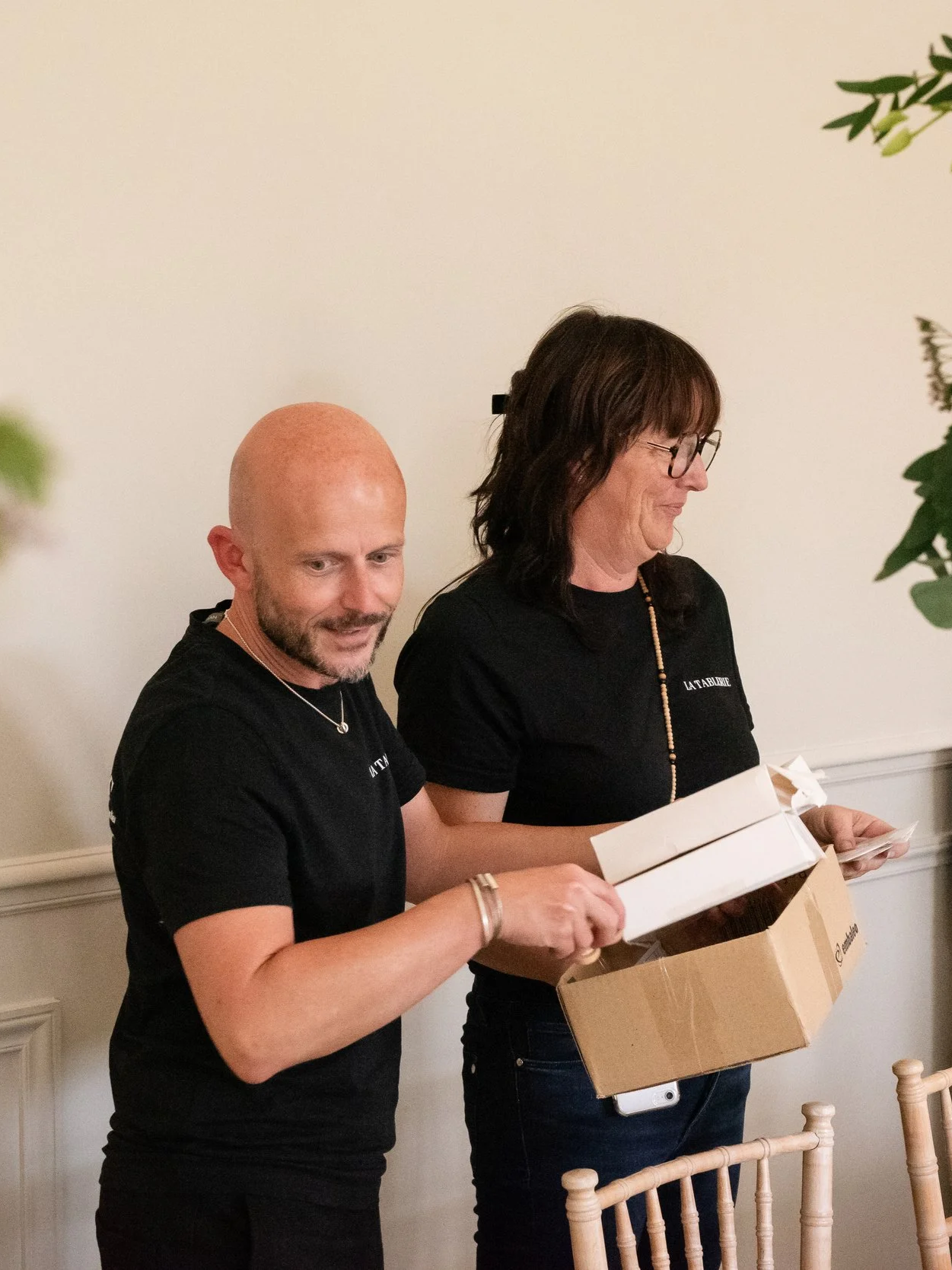 Deux personnes, un homme chauve avec une barbe et une femme avec des lunettes, ouvrent un carton avec du contenu à l'intérieur dans une pièce intérieure. Ils portent tous deux un t-shirt noir.