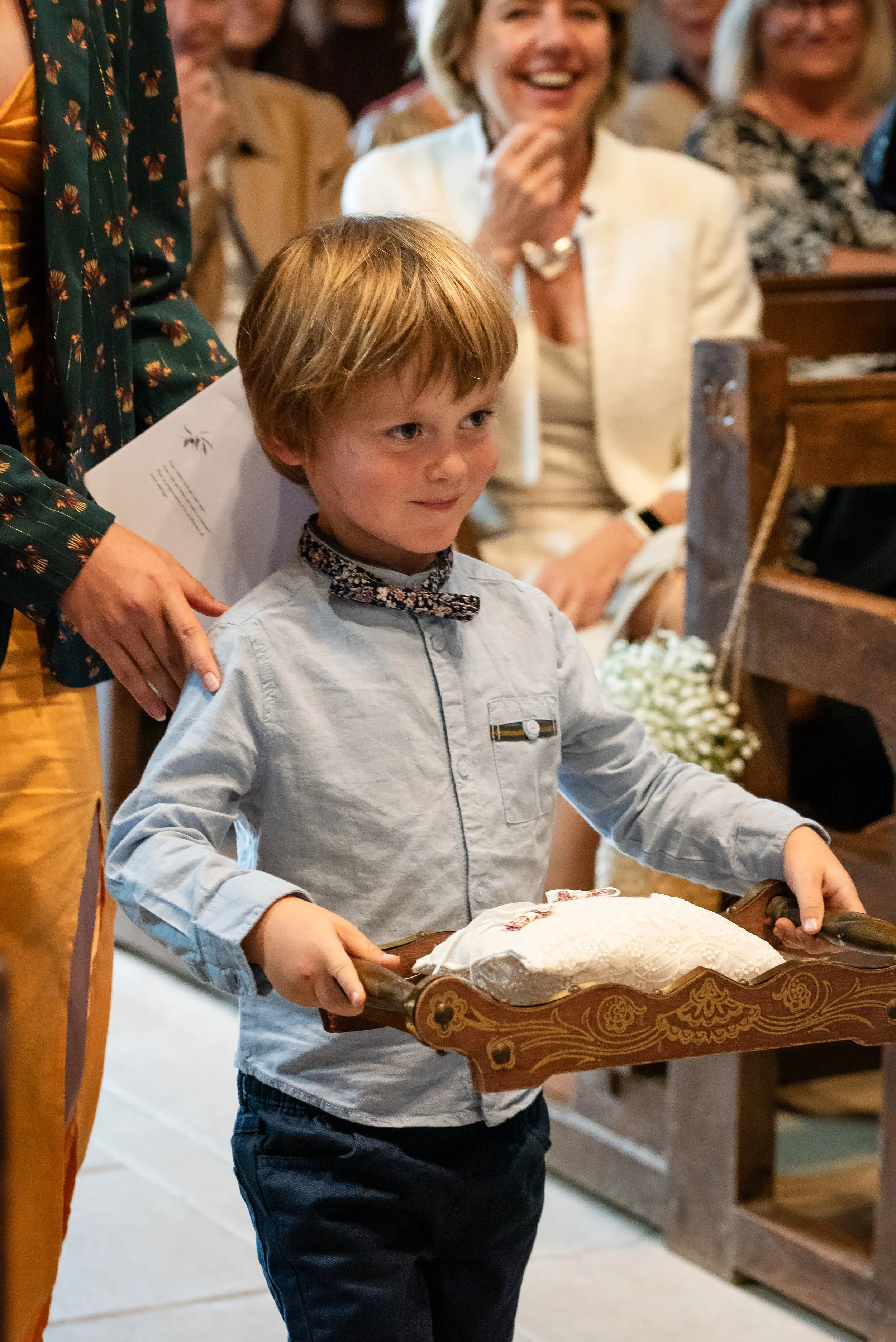 Un garçon tient un plateau en bois avec un gâteau recouvert de sucre glace lors d'une cérémonie, entouré de personnes applaudissant et souriant dans un intérieur chaleureusement décoré.