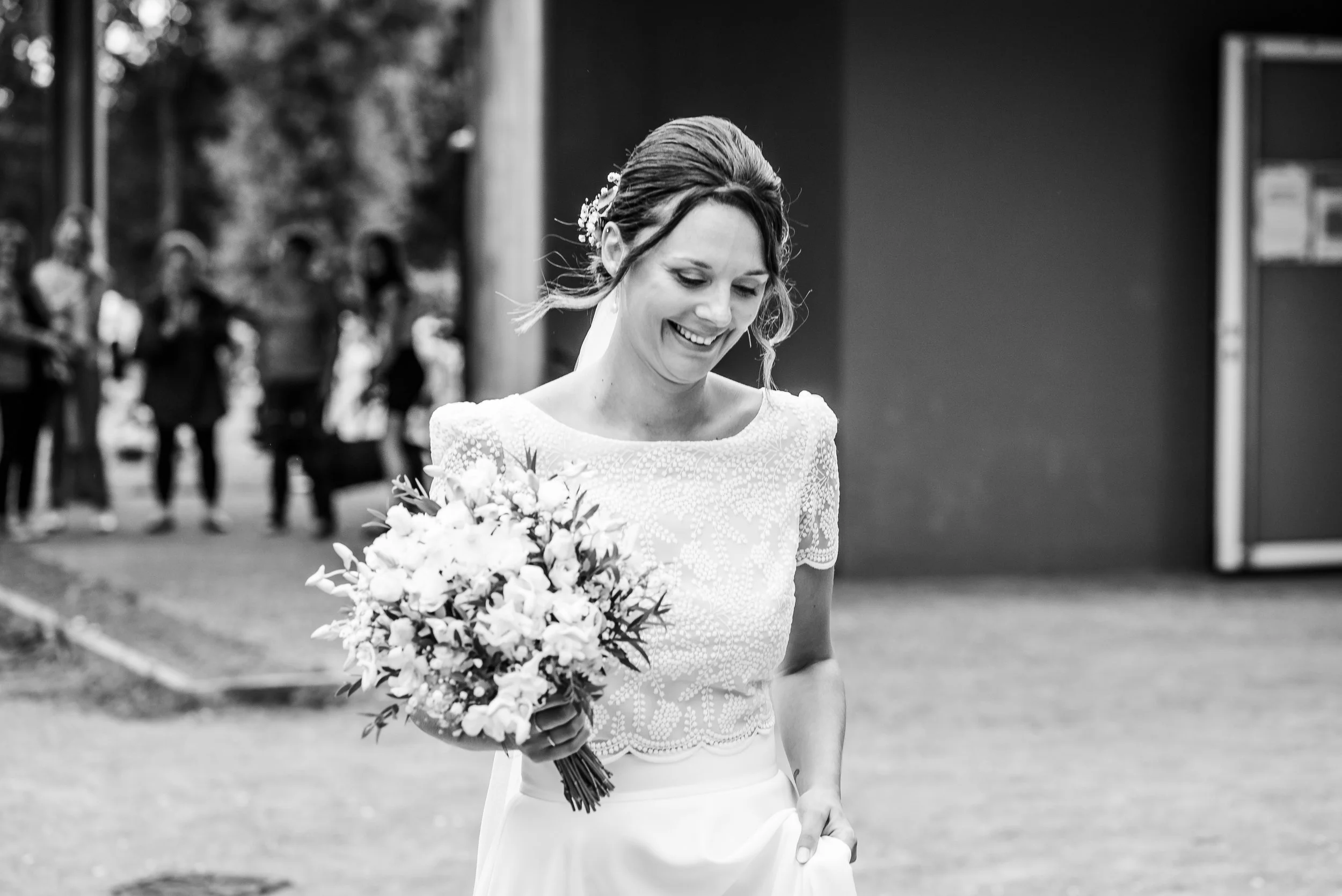 Une femme souriante en robe de mariée portant un bouquet de fleurs lors d'une cérémonie de mariage en plein air.