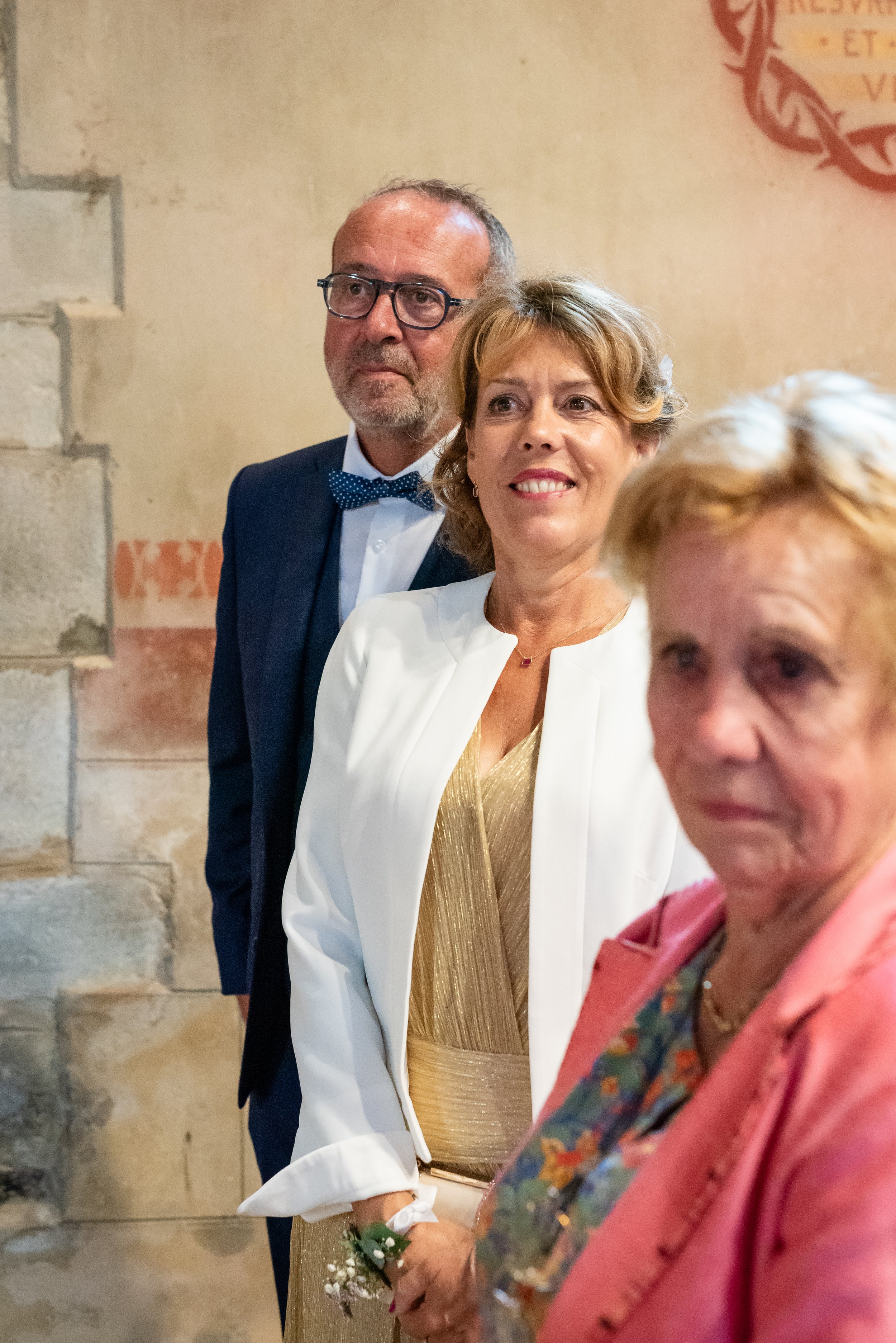 Trois personnes à une cérémonie, deux femmes et un homme, en tenue formelle, dans un lieu avec un mur en pierre. La femme au centre porte une robe dorée et un blazer blanc, la femme à droite porte un blazer rose avec un foulard à motifs colorés, l'ho