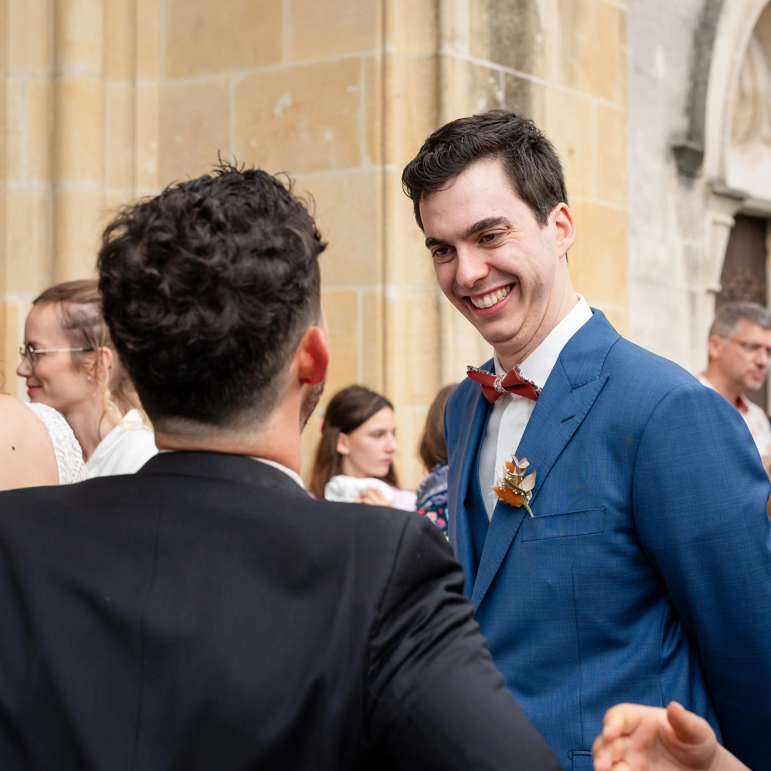Jeune homme portant un costume bleu avec un nœud papillon à motifs, souriant, lors d'un événement social dans un lieu avec des murs en pierre.