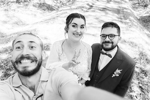 Trois personnes prennent un selfie en extérieur, dont une femme en robe blanche et deux hommes en costumes, tous souriants.