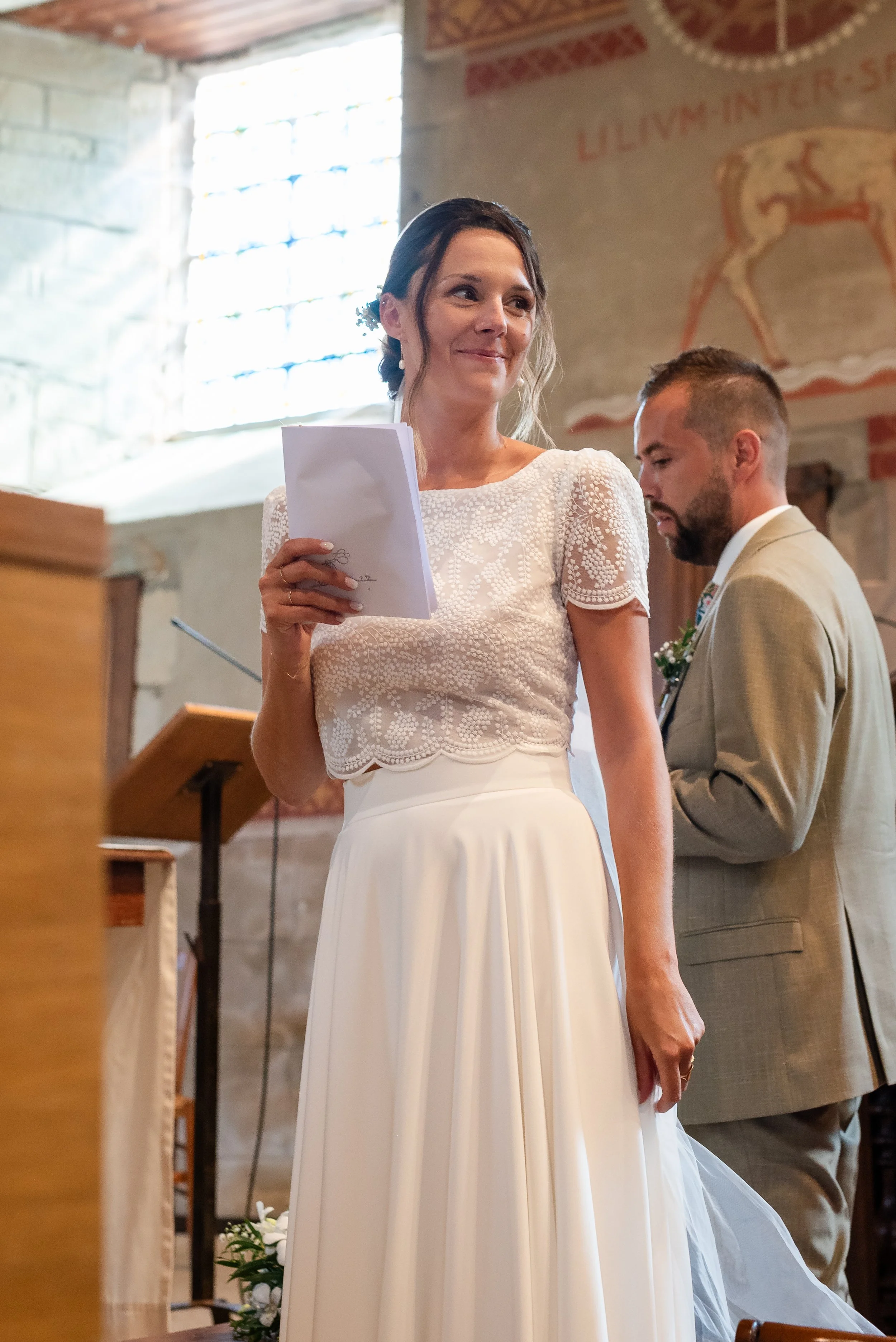 Une femme en robe de mariage tient une feuille, un homme avec un costume beige est à côté lors d'une cérémonie dans une église en pierre avec fenêtres en verre coloré.