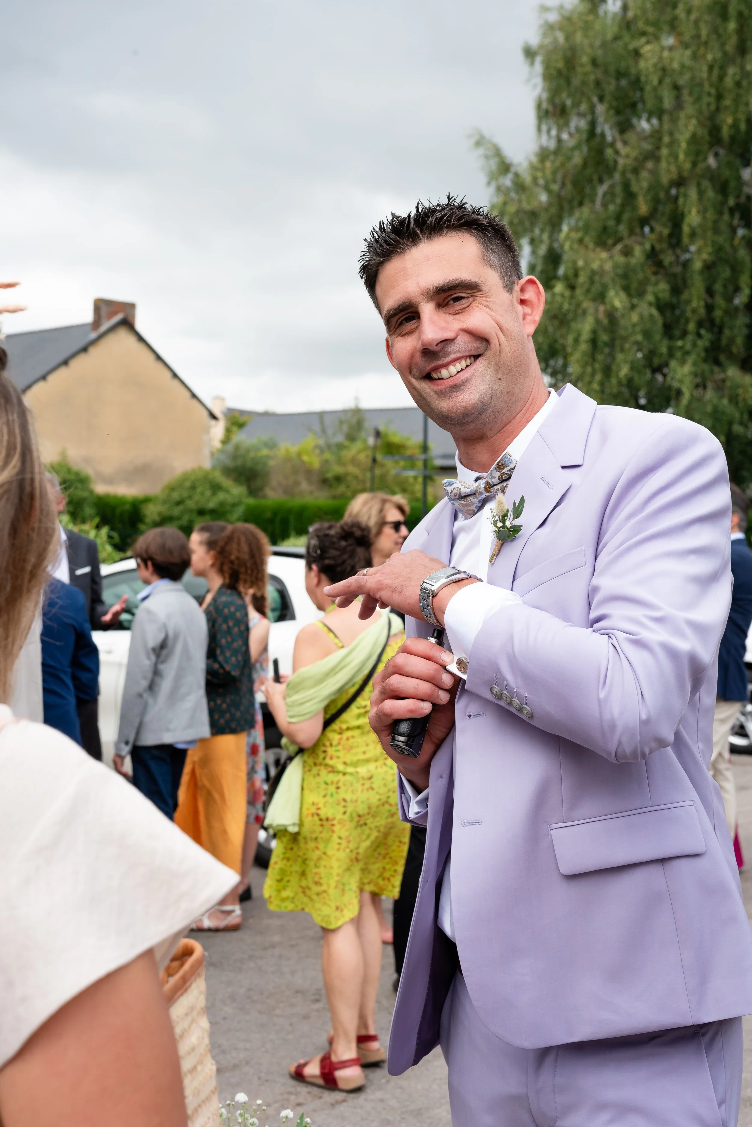 Un homme souriant habillé en costume violet lors d'un événement en plein air, entouré de plusieurs personnes.