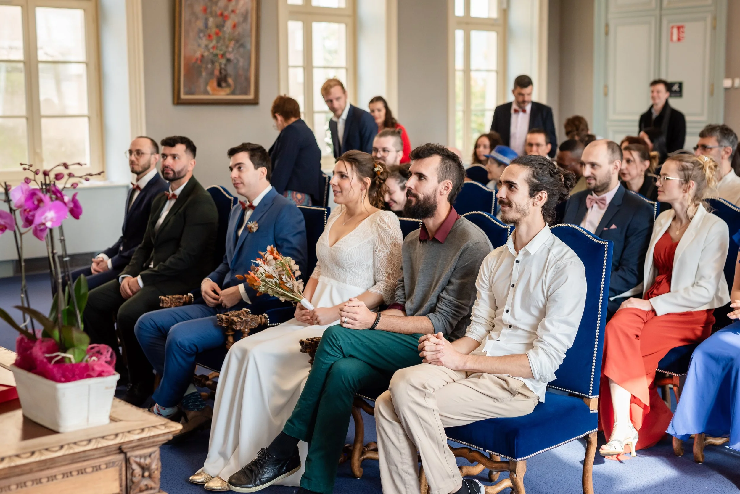 Groupe d'invités assis dans une salle lumineuse lors d'une cérémonie, certains portant des costumes et des robes, avec des fleurs et des décorations autour.