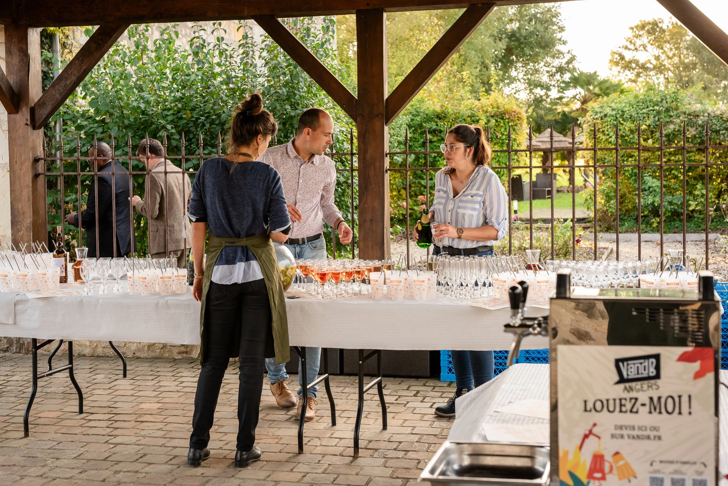 Groupe de personnes conversant devant une table de boissons lors d'un événement en plein air avec un cadre verdoyant.