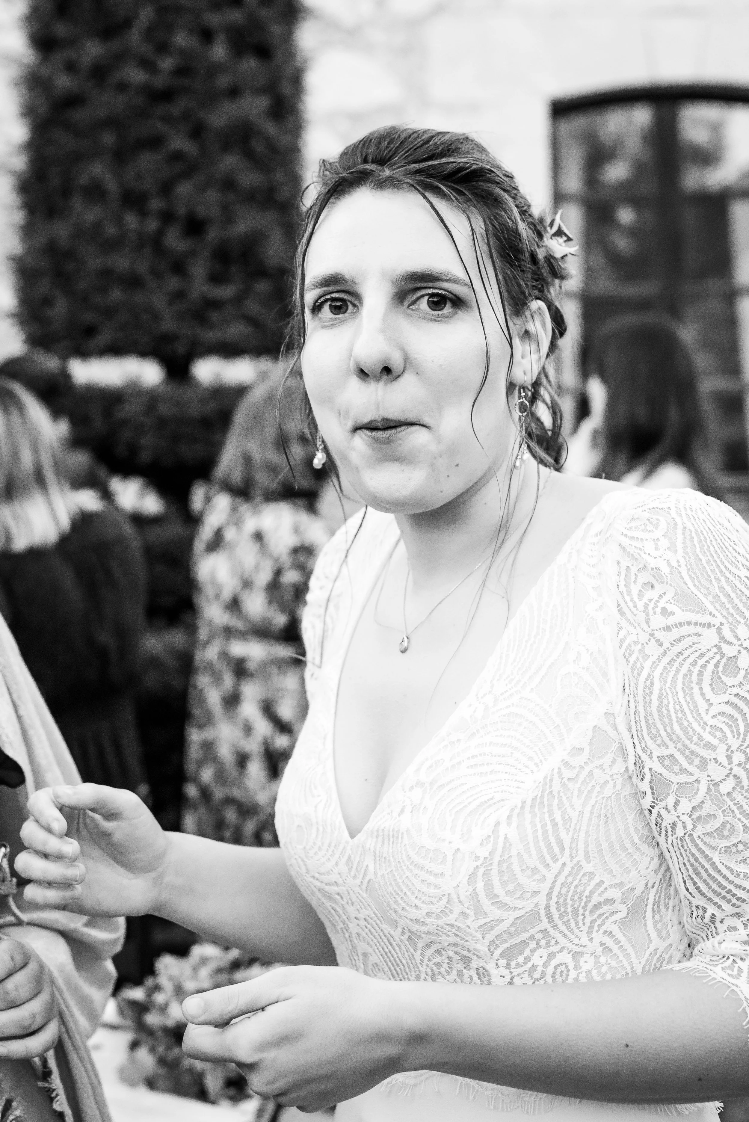 Photo en noir et blanc d'une femme portant une robe en dentelle, avec des cheveux attachés, portant des boucles d'oreilles et un collier, discutant avec d'autres personnes lors d'un événement à l'extérieur.