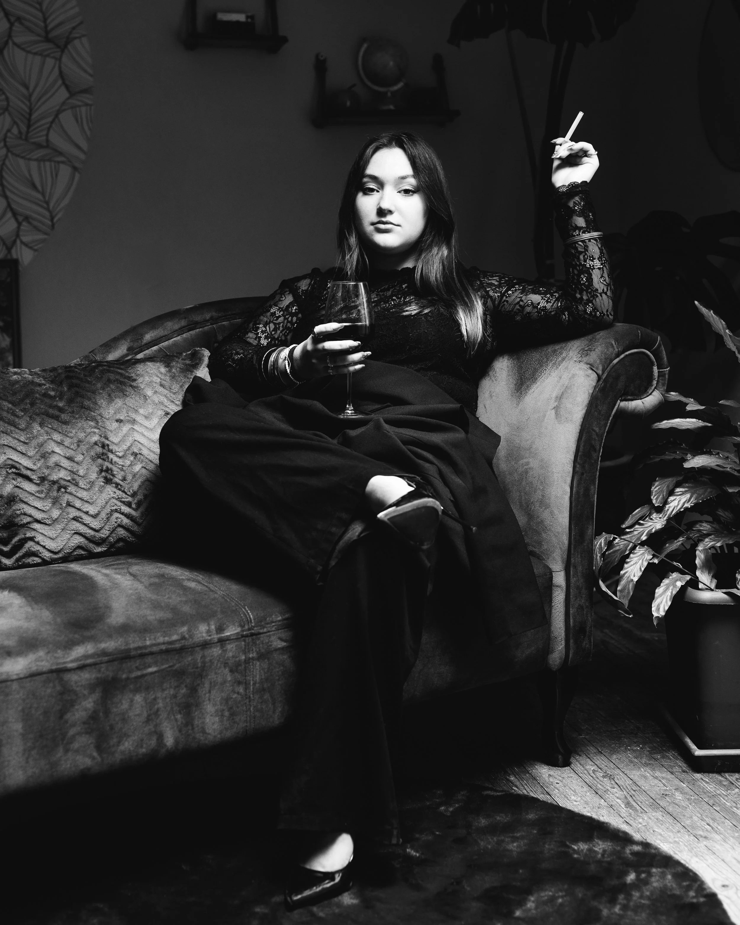 Jeune femme assise sur un canapé, tenant un verre de vin rouge dans une main et un cigarette dans l'autre, vêtue d'une robe en dentelle noire, avec un décor intérieur, incluant une étagère avec un globe terrestre en arrière-plan.