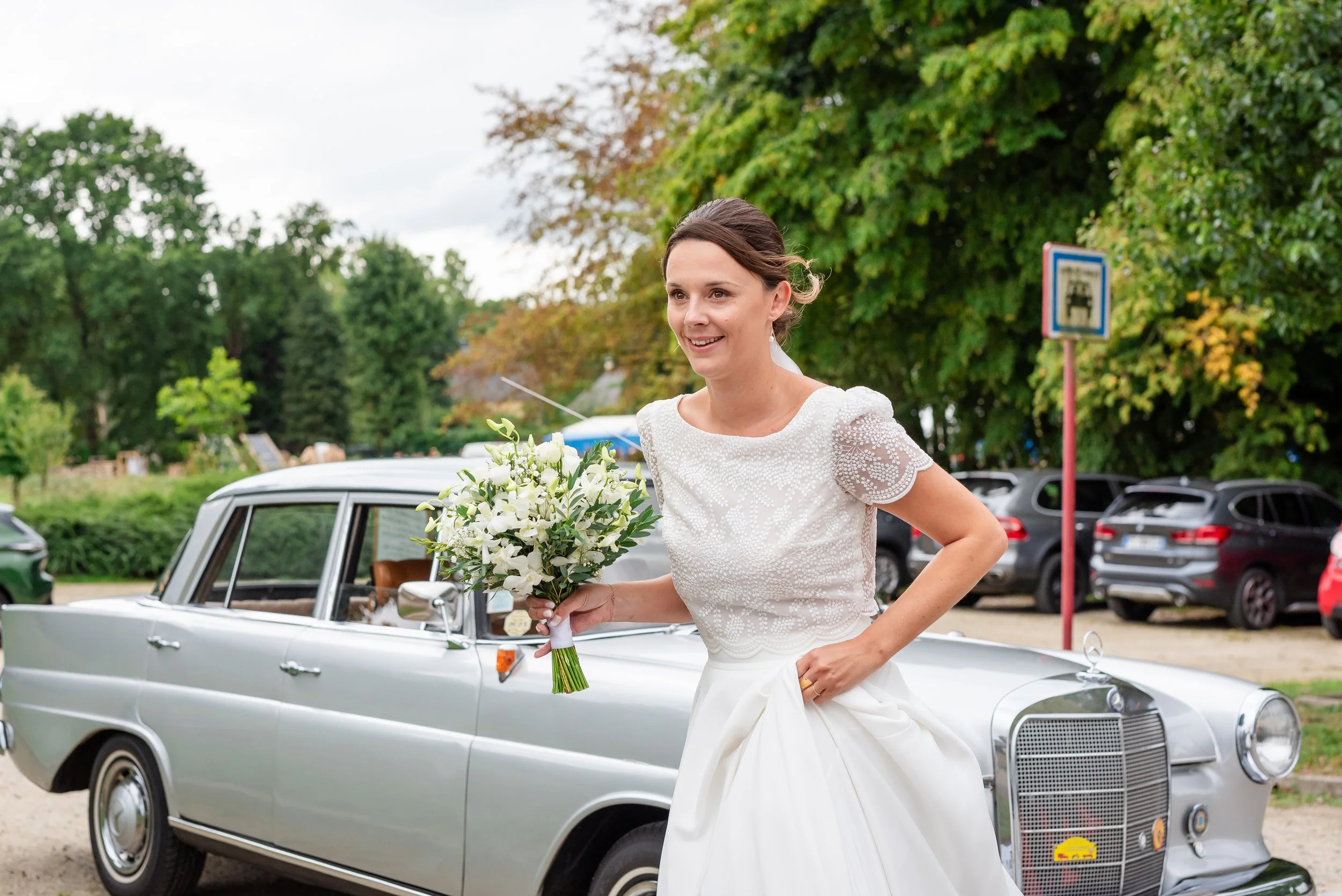 Une femme en robe blanche de mariage tient un bouquet de fleurs blanches et sourit, debout devant une voiture classique argentée, avec des arbres feuillus en arrière-plan.