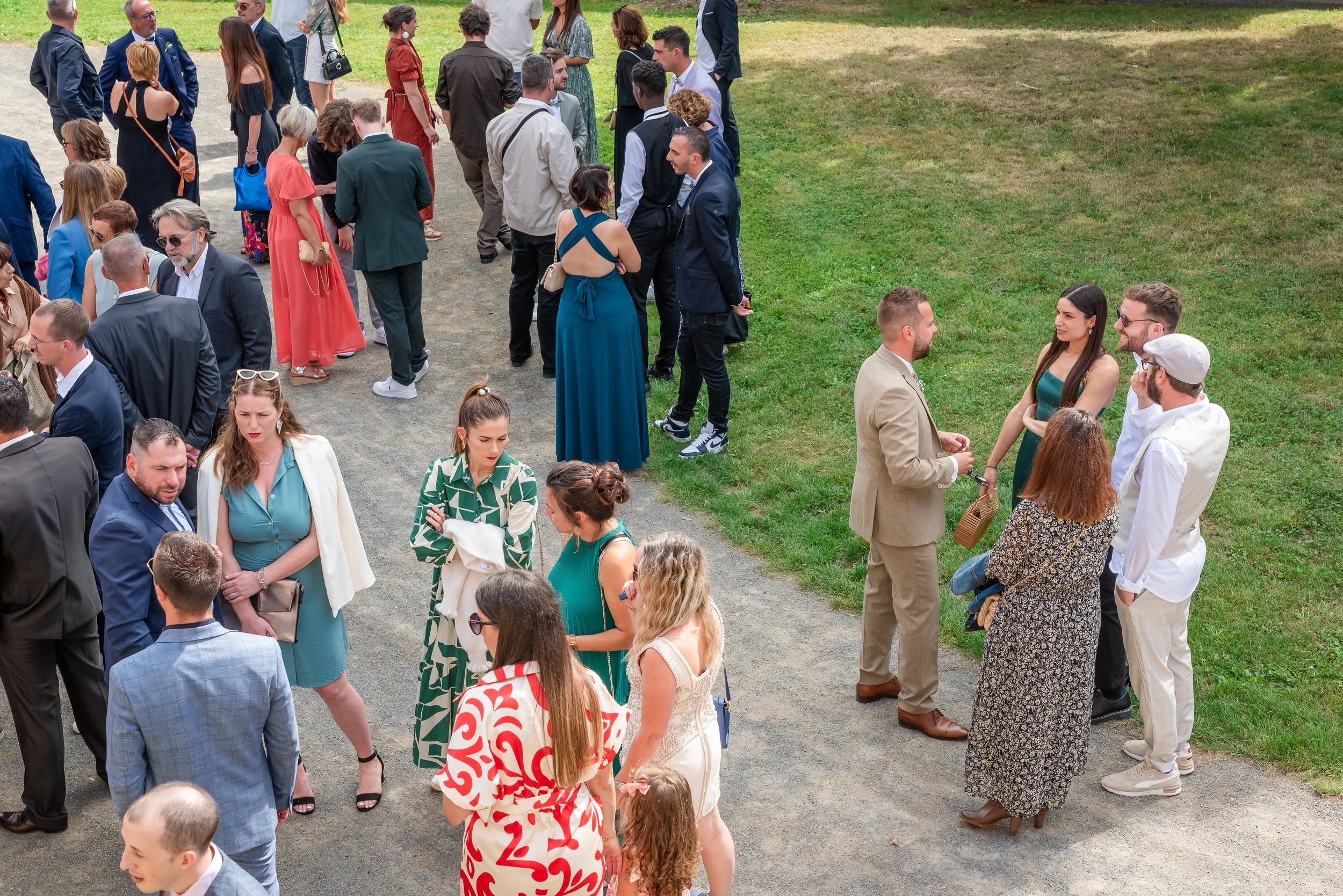 Groupe de personnes habillées en vêtements formels, probablement lors d'un mariage ou d'une cérémonie en plein air, dû à une disposition en ligne et le cadre extérieur avec de l'herbe et un chemin.