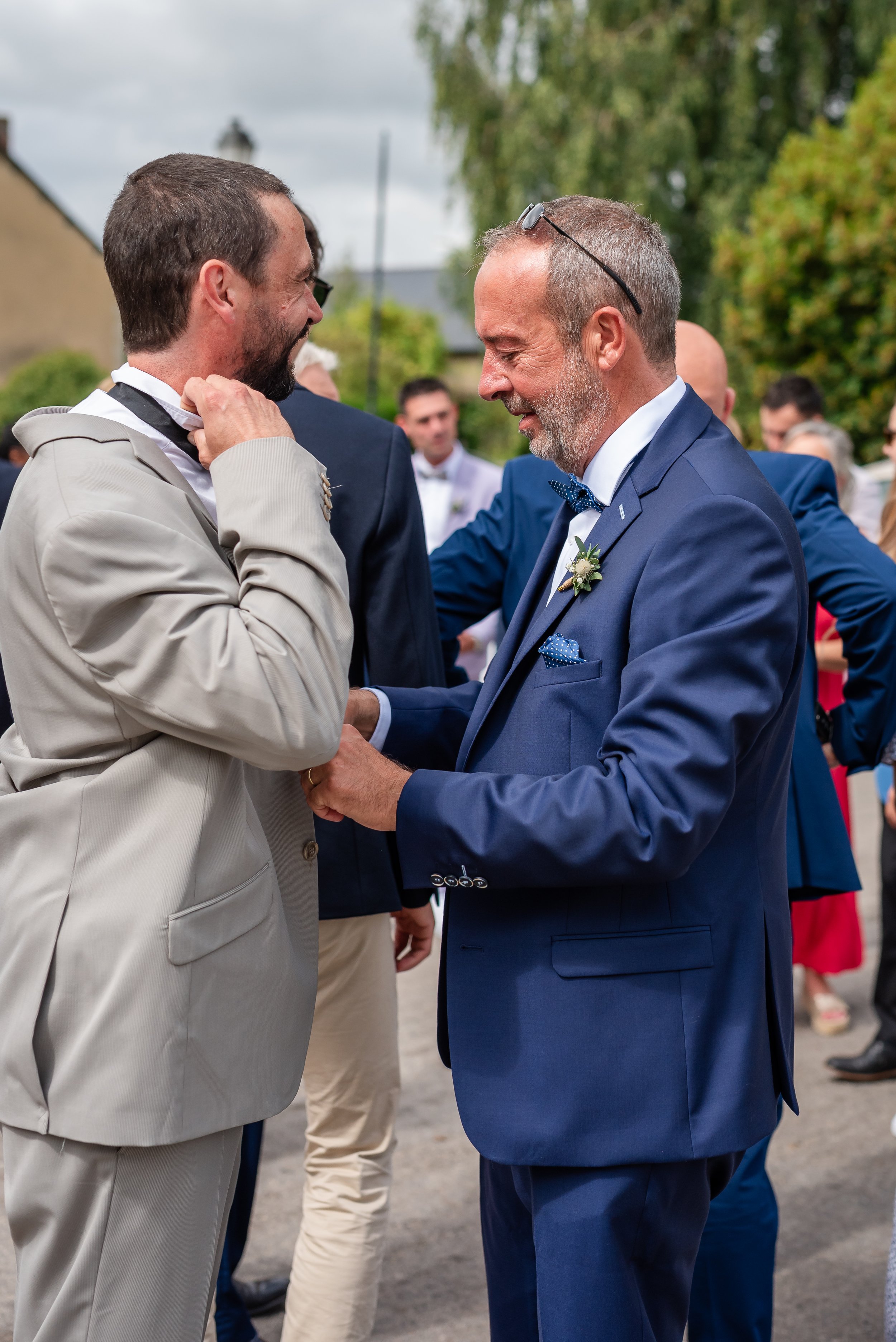 Deux hommes habillés en costume lors d'un mariage, échangeant un sourire et se tenant la main dans un espace extérieur avec des invités en arrière-plan.