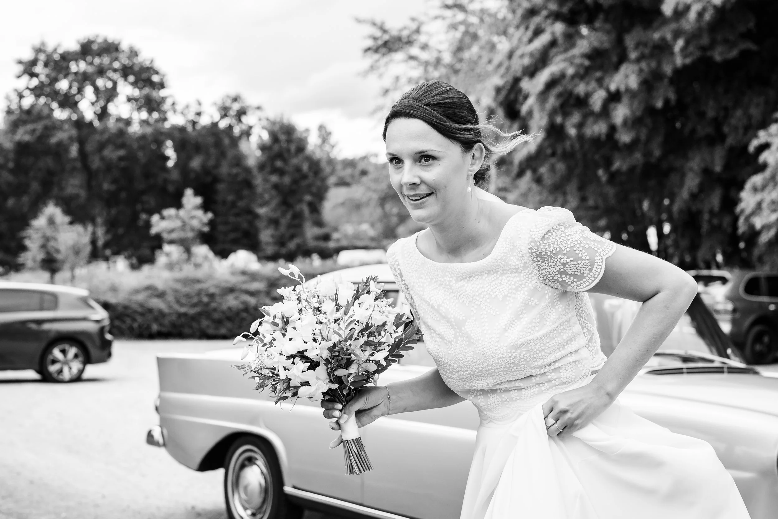 Femme souriante en robe de mariage tenant un bouquet de fleurs, debout à côté d'une voiture, en extérieur, en noir et blanc.