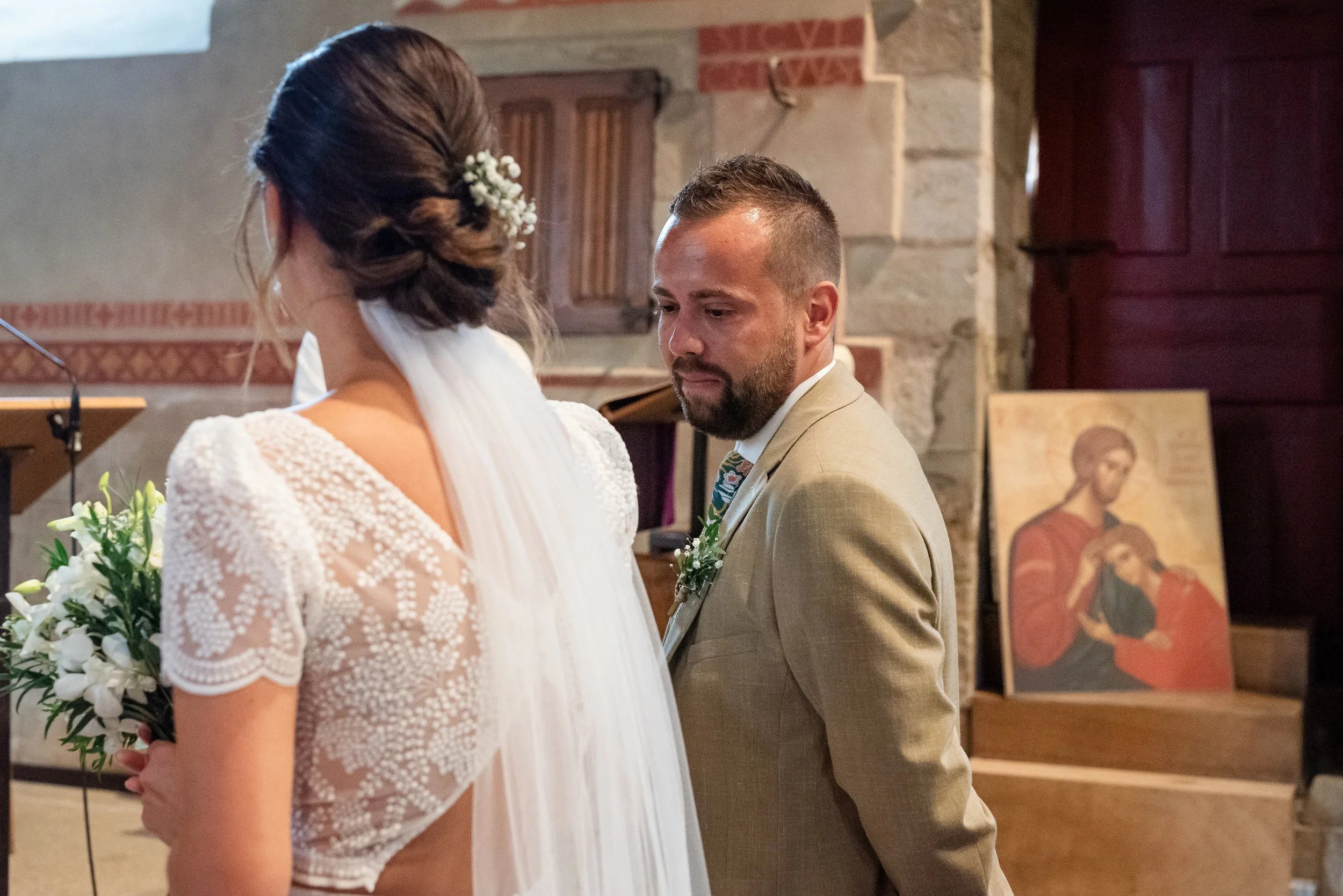 Un couple lors d'une cérémonie de mariage religieuse, l'homme en costume beige et la femme en robe blanche, dans une église en pierre, avec des icônes religieuses en arrière-plan.