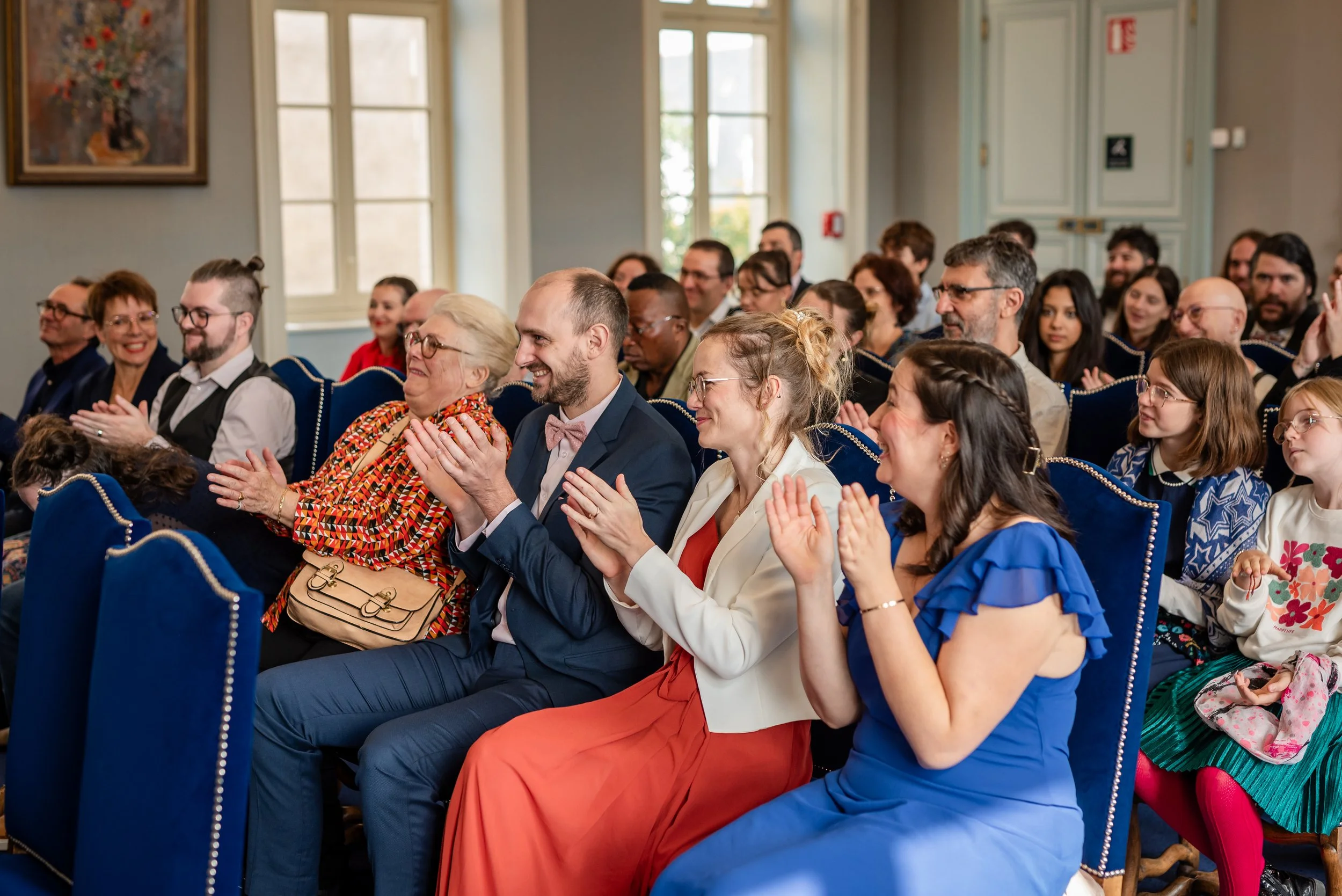 Un groupe de personnes assises dans une salle, applaudissant et souriant lors d'un événement ou d'une cérémonie. La salle est lumineuse avec de grandes fenêtres et des meubles en bois, décorée avec un tableau floral sur le mur.