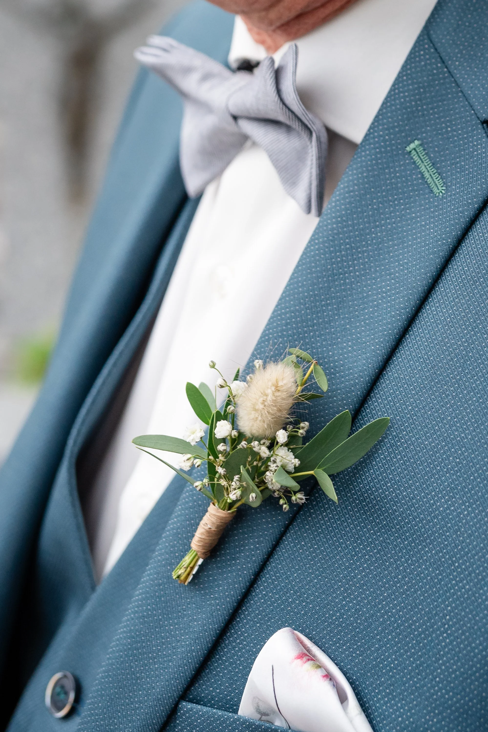 Un homme en costume bleu avec un nœud papillon gris porte une boutonnière avec des fleurs blanches, des feuilles vertes et une touffe de coton. Il y a également une pochette dans la poche de sa veste.