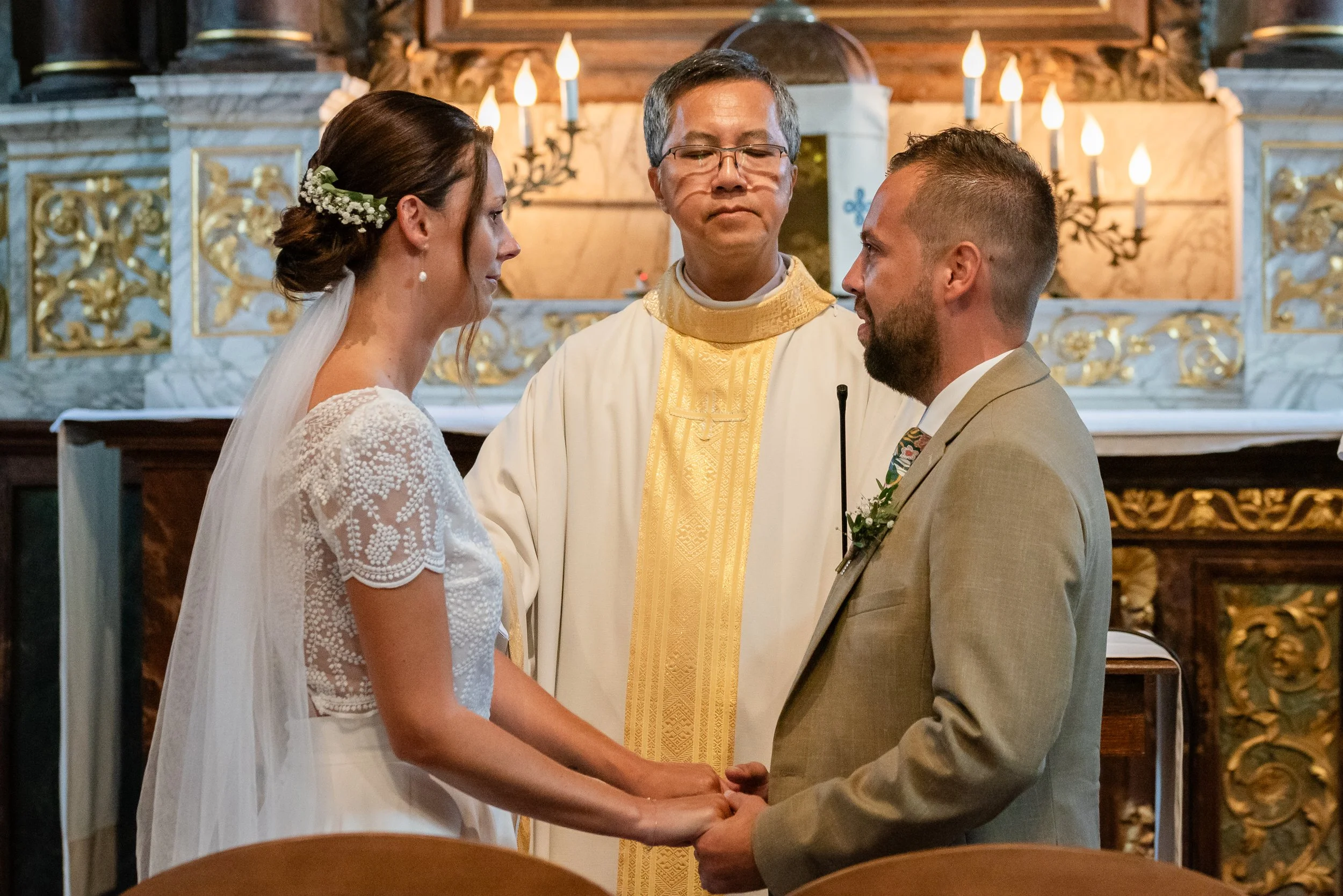 Un couple se tenant par la main lors de leur mariage dans une église, face à un prêtre. La mariée porte une robe blanche à manches en dentelle avec une coiffure ornée de fleurs, et le marié porte un costume beige.