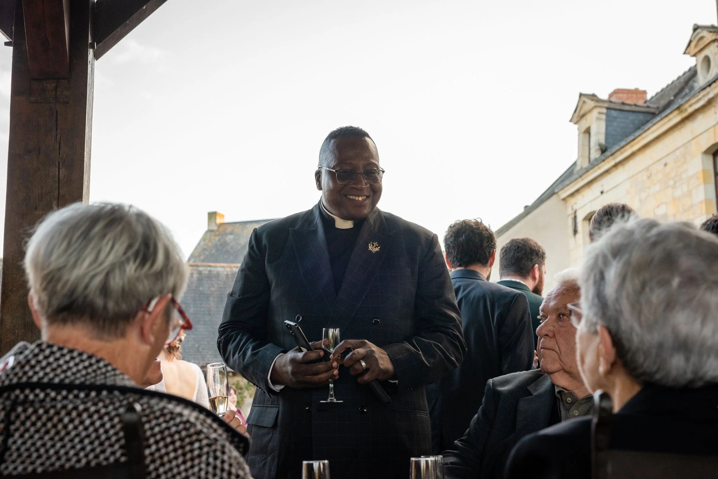 Un homme en habit d'homme d'église parlant à un groupe de personnes âgées lors d'un événement en plein air avec des bâtiments en fond.