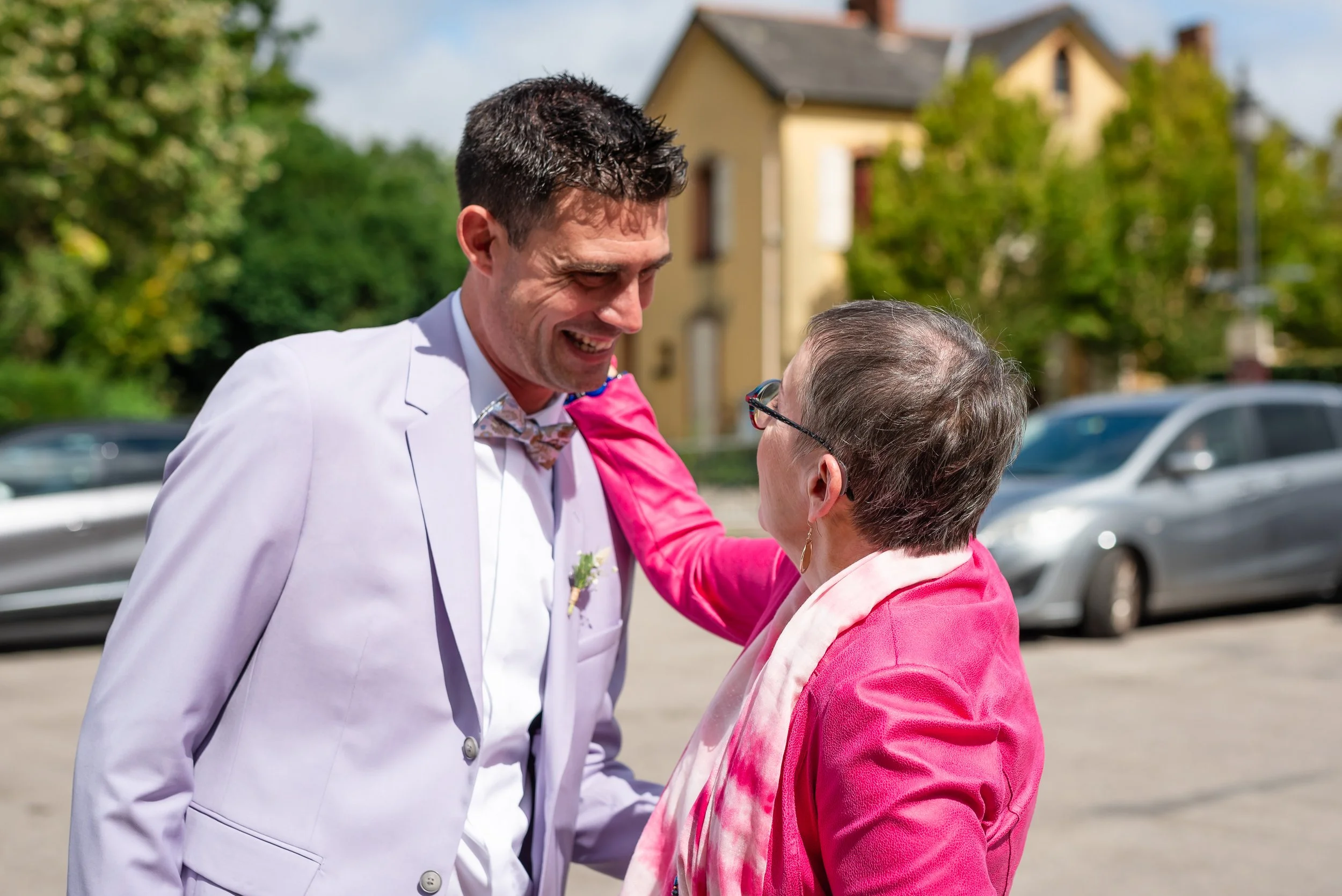 Un homme en costume blanc souriant et une femme en rouge se saluant à l'extérieur devant une maison et des voitures.
