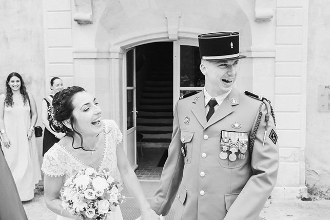 Une mariée en robe de mariage souriant et tenant un bouquet, un officier en uniforme militaire également souriant, avec deux autres femmes en robe en arrière-plan, devant un bâtiment en pierre.