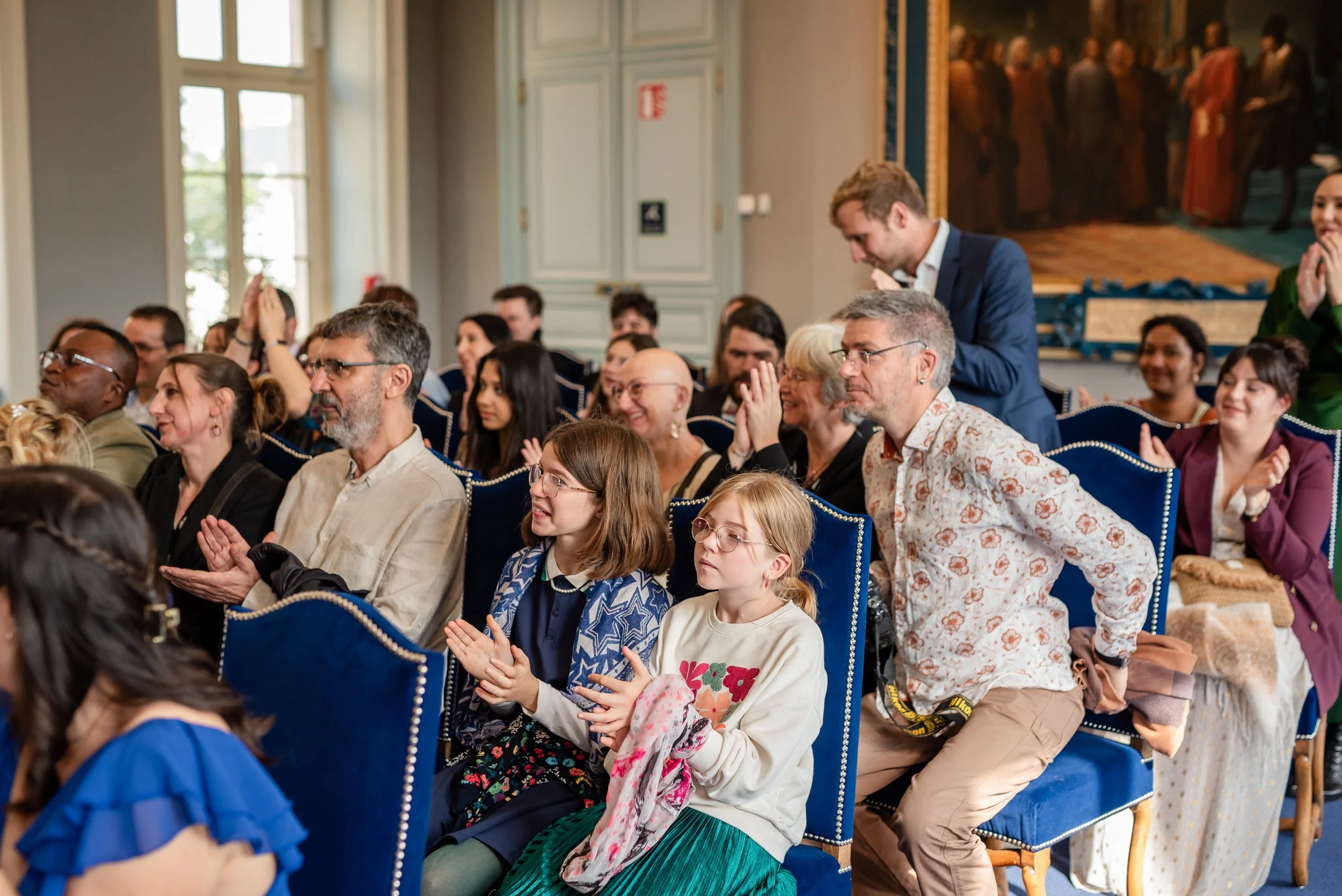 Un groupe de personnes assises en réunion ou en conférence dans une salle ornée, certaines applaudissant et d'autres discutant.