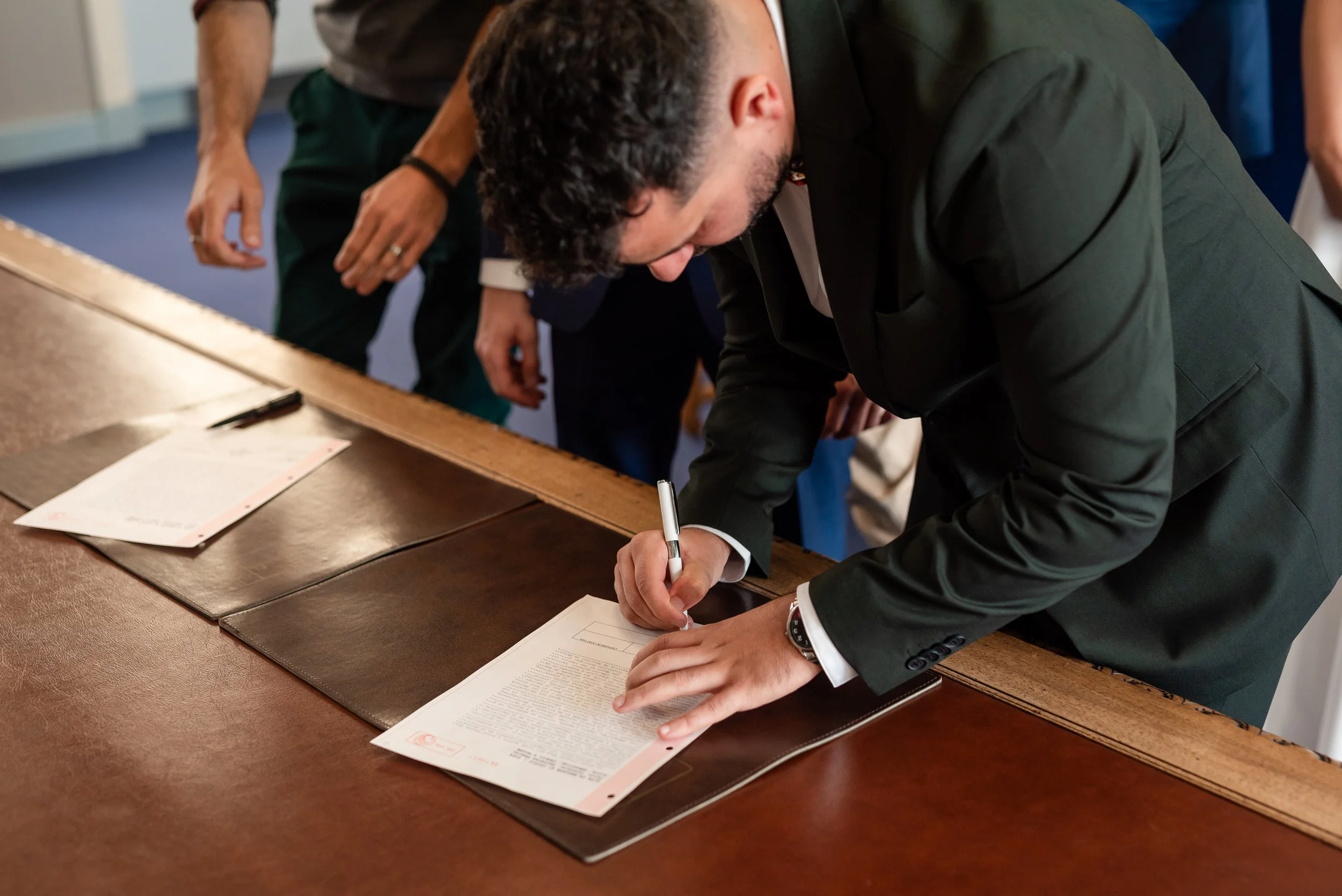 Une personne en costume signe un document posé sur une table en bois, entourée d'autres personnes. La scène se déroule probablement lors d'une cérémonie officielle ou d'une réunion importante.