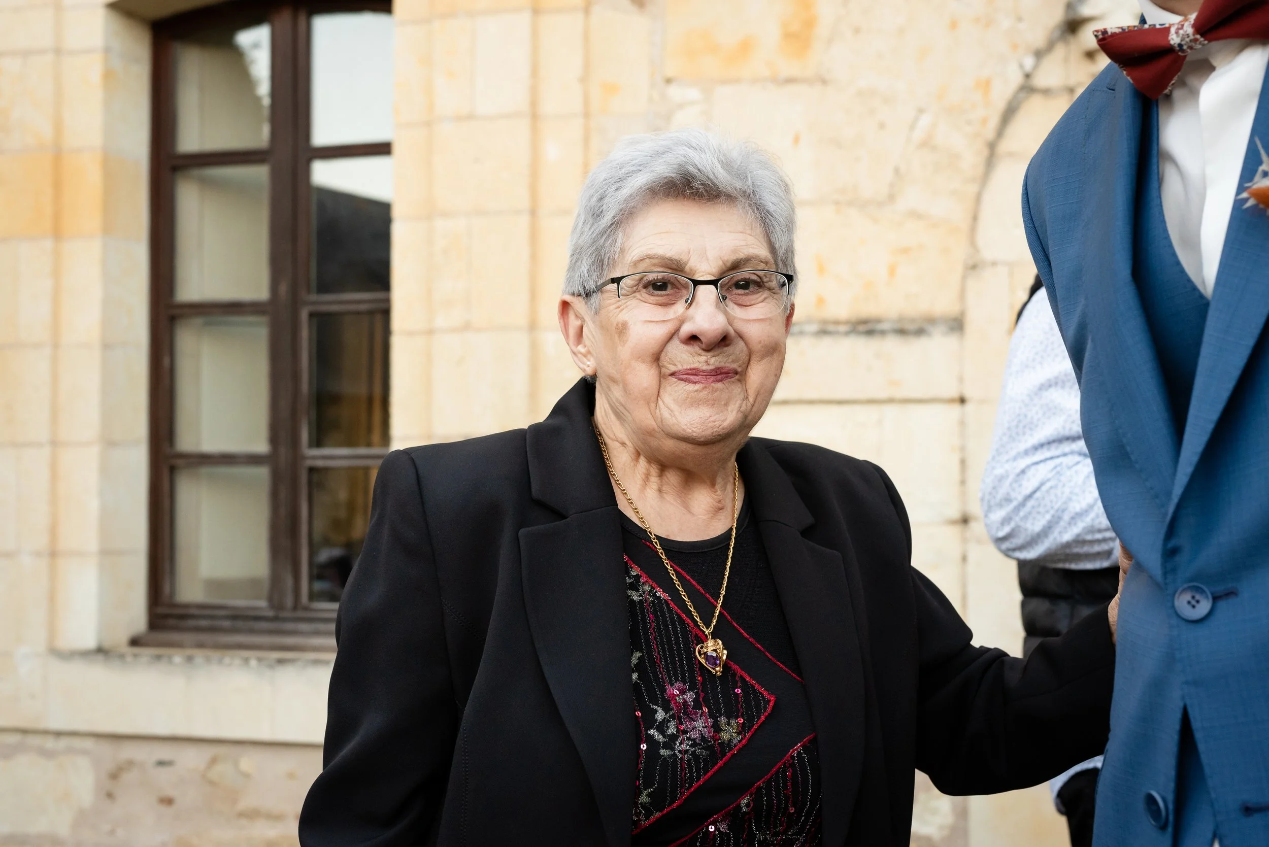 Une femme âgée souriante, portant des lunettes, habillée en noir avec un collier ornemental, serrant la main d'une personne en costume bleu, en extérieur devant un bâtiment en pierre.