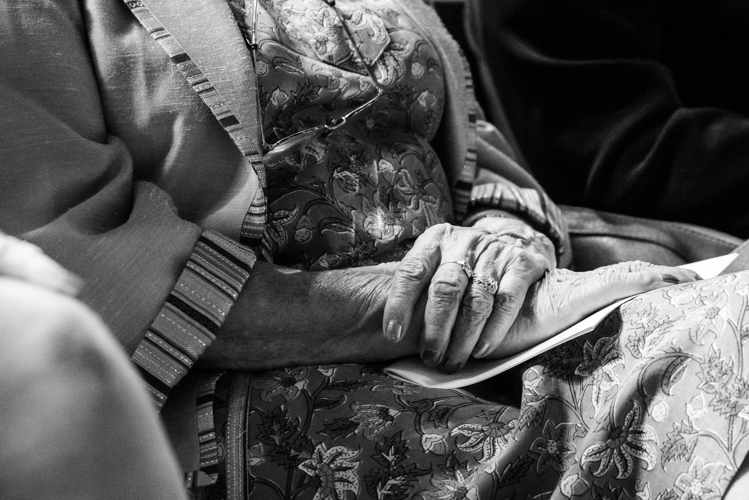 Mains d'une personne âgée reposant sur un livre ou un journal, tenant une paire de lunettes. La photo est en noir et blanc, et la personne porte des vêtements aux motifs floraux et à rayures.
