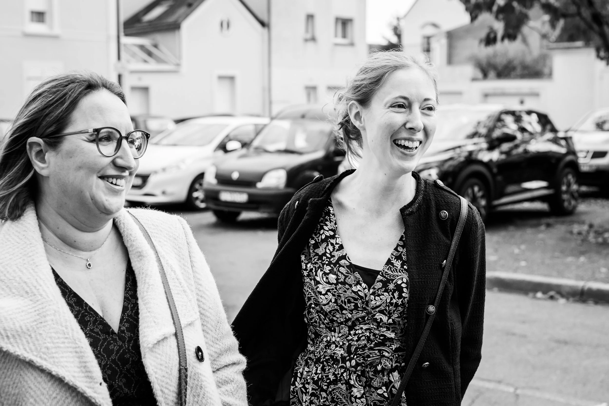 Deux femmes souriantes marchant côte à côte dans une zone urbaine, avec des voitures garées en arrière-plan.