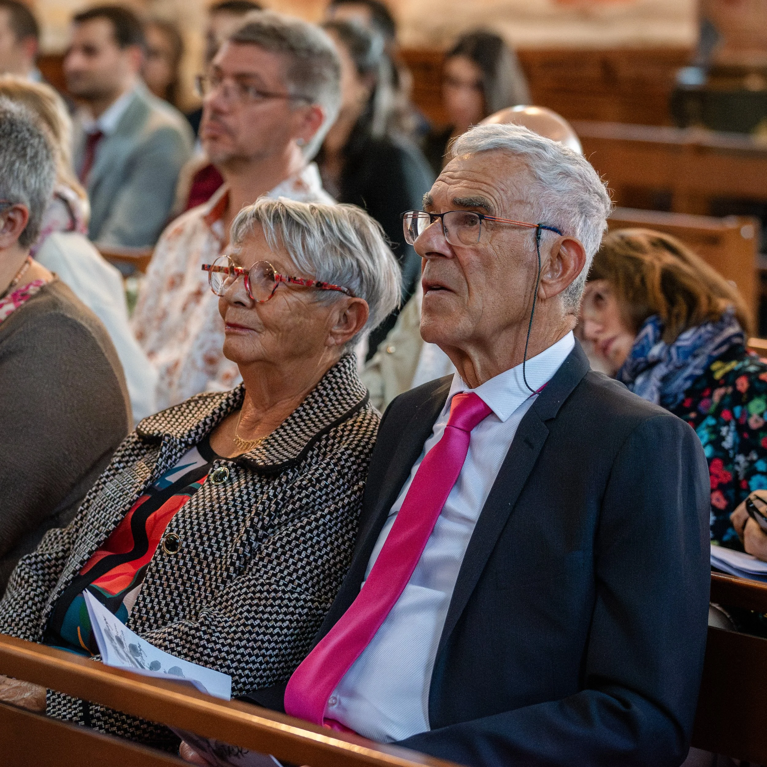 Groupe de personnes assises en réunion, la plupart portent des lunettes et sont concentrées sur un événement ou un discours, dans une salle avec bancs en bois.
