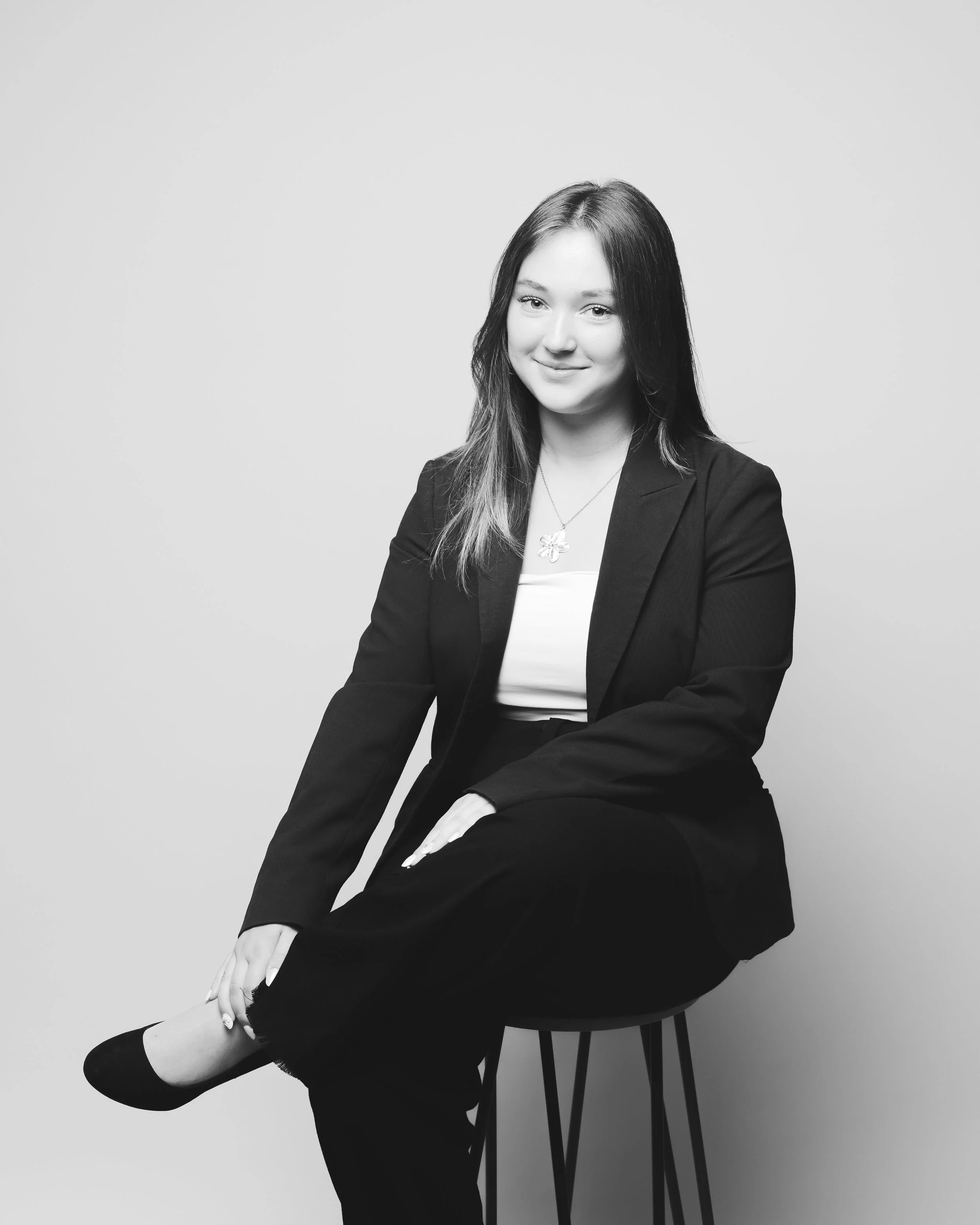 Portrait d'une jeune femme souriante, portant un blazer noir, assise sur une chaise, en noir et blanc.