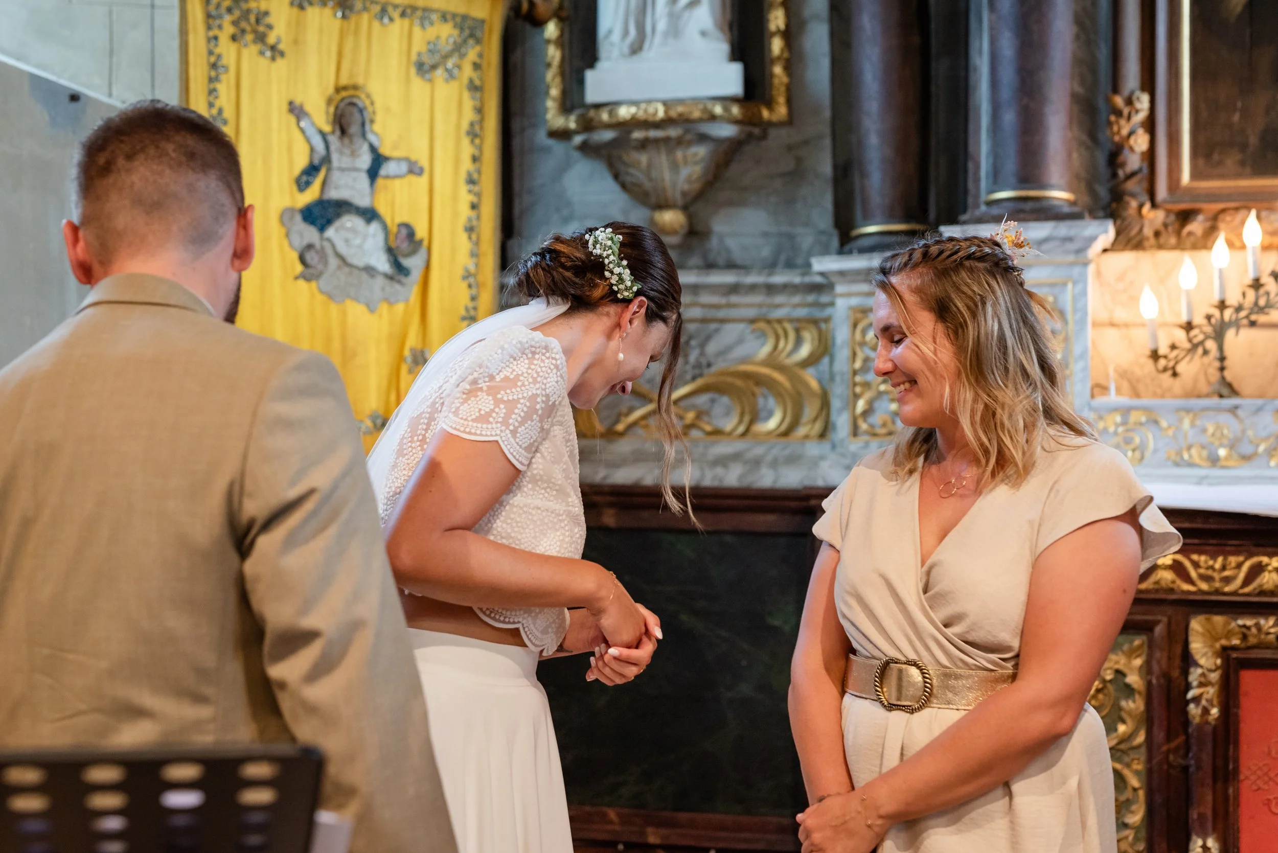 Une scène de mariage à l'intérieur d'une église avec quatre personnes : un couple au centre, une femme à droite, et un homme à gauche dont le dos est visible. La femme se trouve devant le couple, souriante, avec des cheveux bouclés, portant une robe 