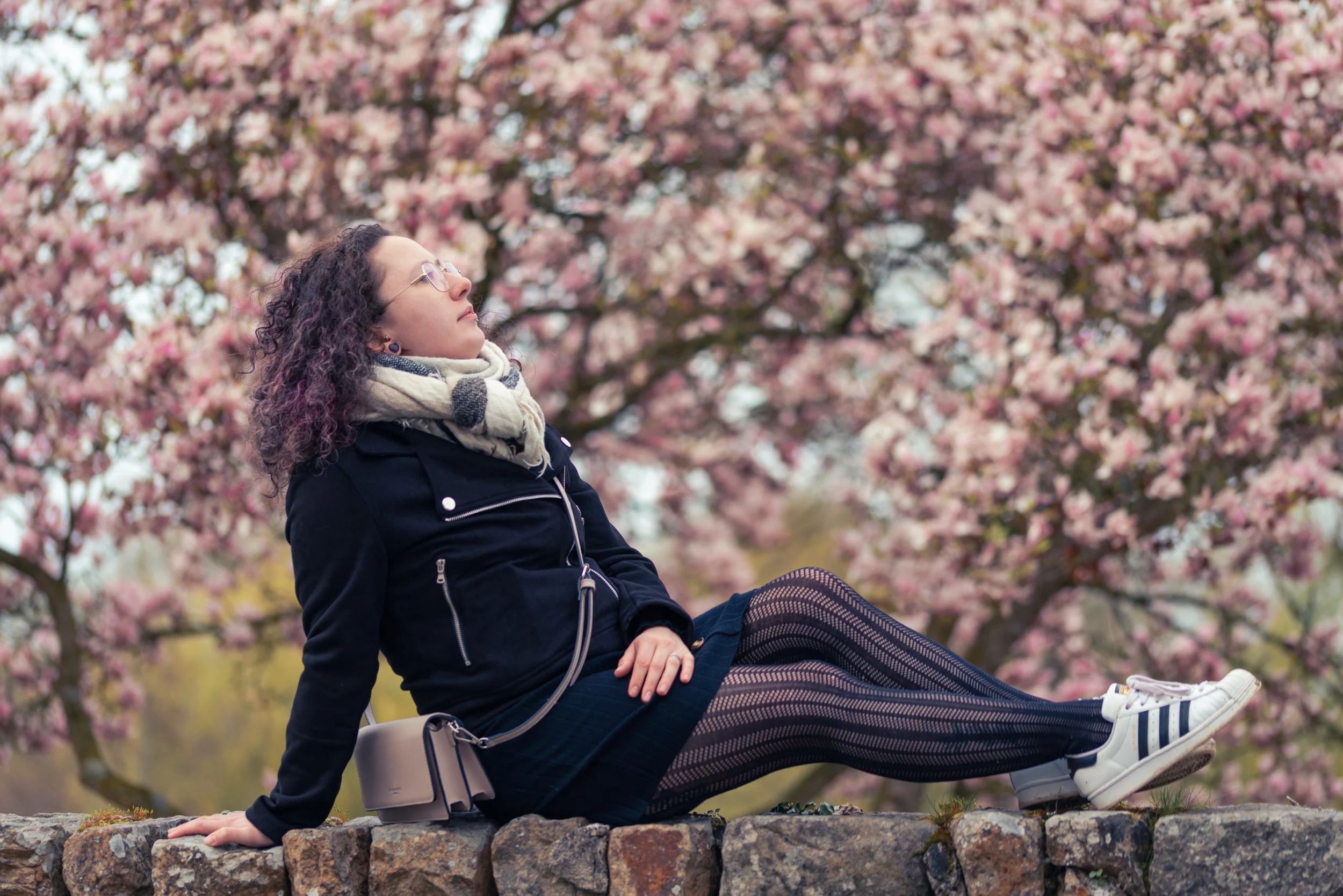 Une femme aux cheveux bouclés portant des lunettes, un manteau noir, une écharpe blanche avec des motifs, assise sur un muret en pierre devant un arbre en fleurs roses. Elle regarde vers le haut, dans une posture détendue.
