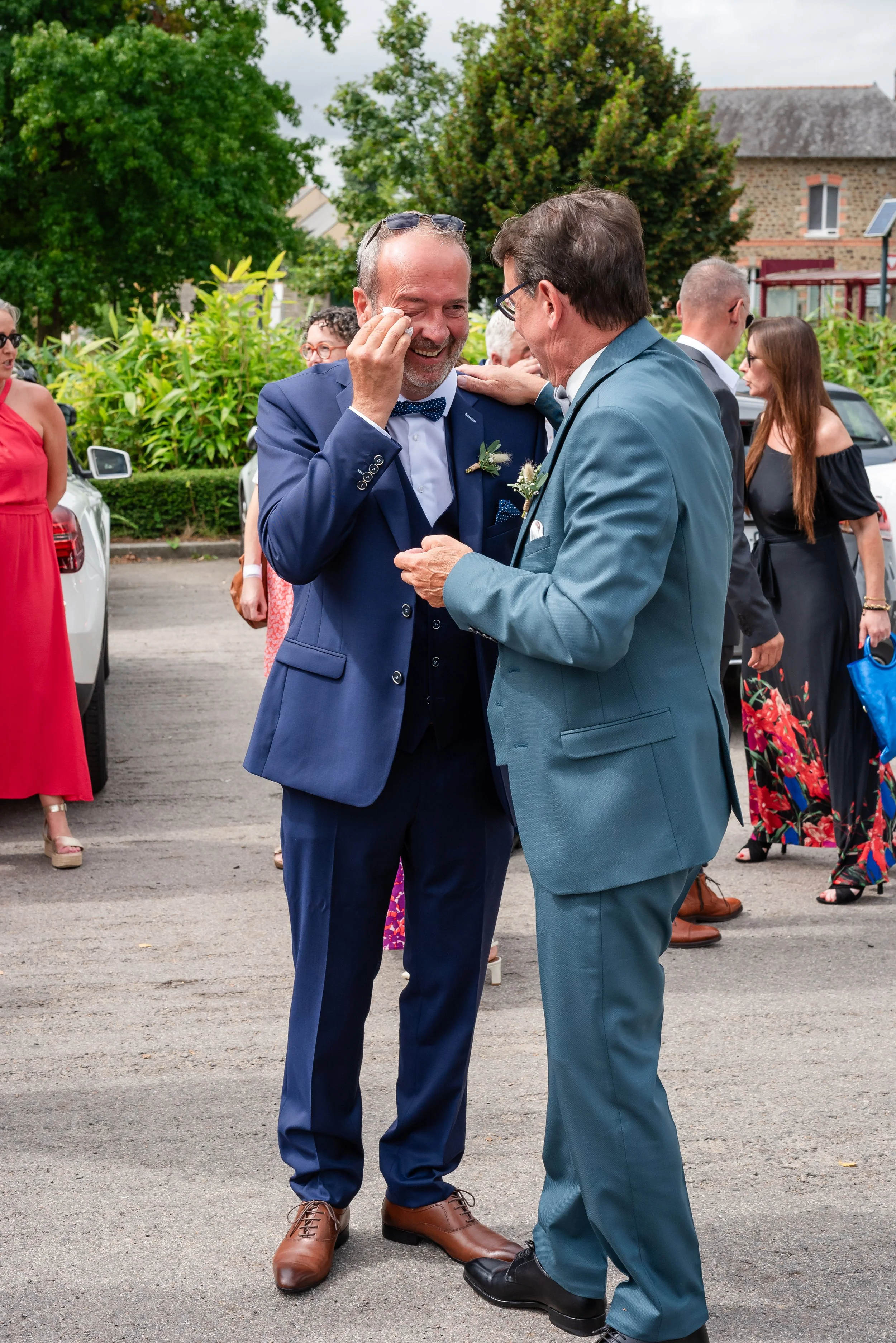 Deux hommes en costume, dont l'un essuie une larme de joie, partagent un moment d'émotion lors d'un mariage en plein air. Autour d'eux, plusieurs personnes en tenues élégantes assistent à l'événement, dans un décor verdoyant avec des arbres et une vi