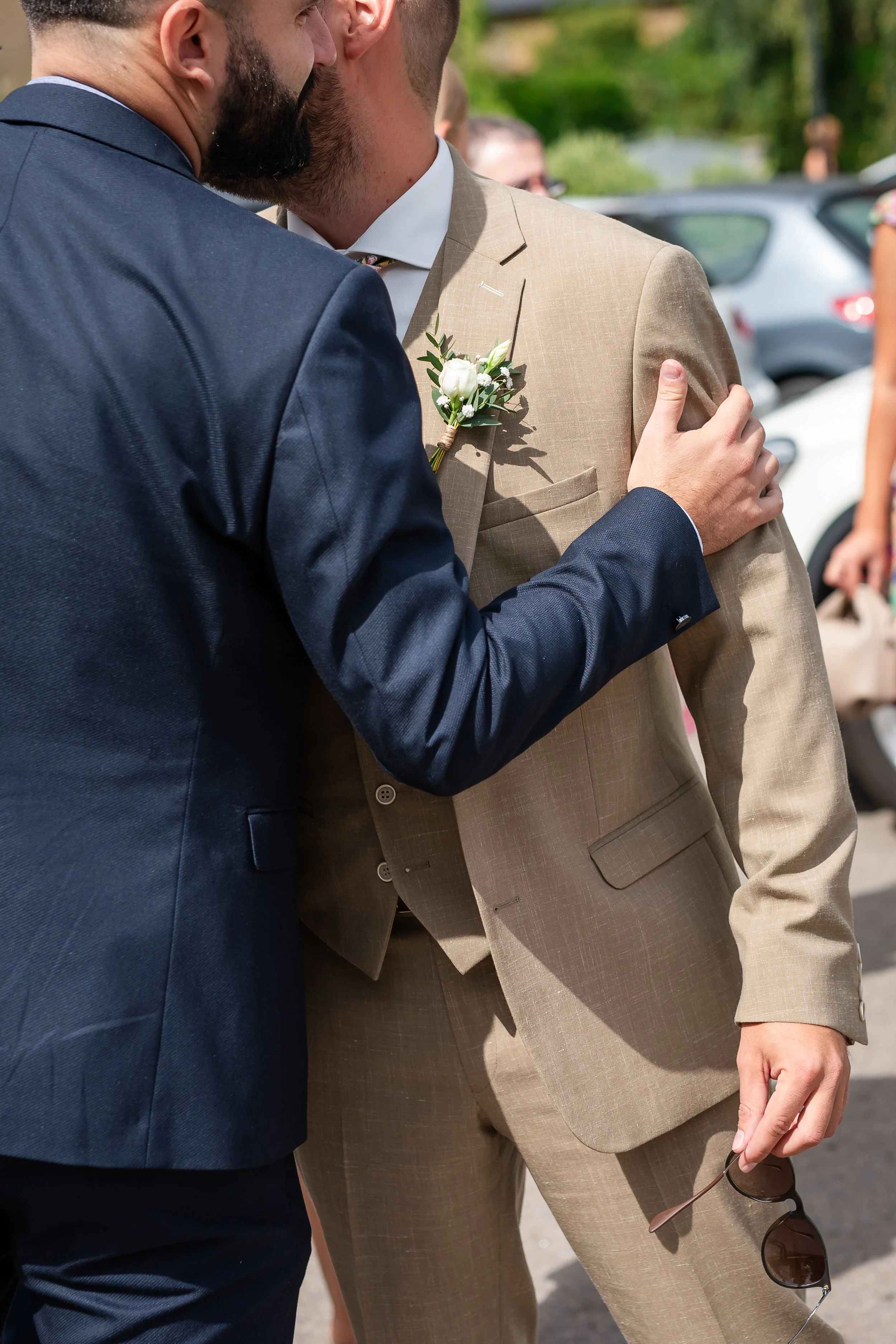 Deux hommes habillés en costumes discutent lors d’un événement en plein air. L’un porte un costume beige avec une boutonnière florale, l’autre un costume bleu foncé. Leurs visages ne sont pas visibles, mais ils semblent engagés dans une conversation 