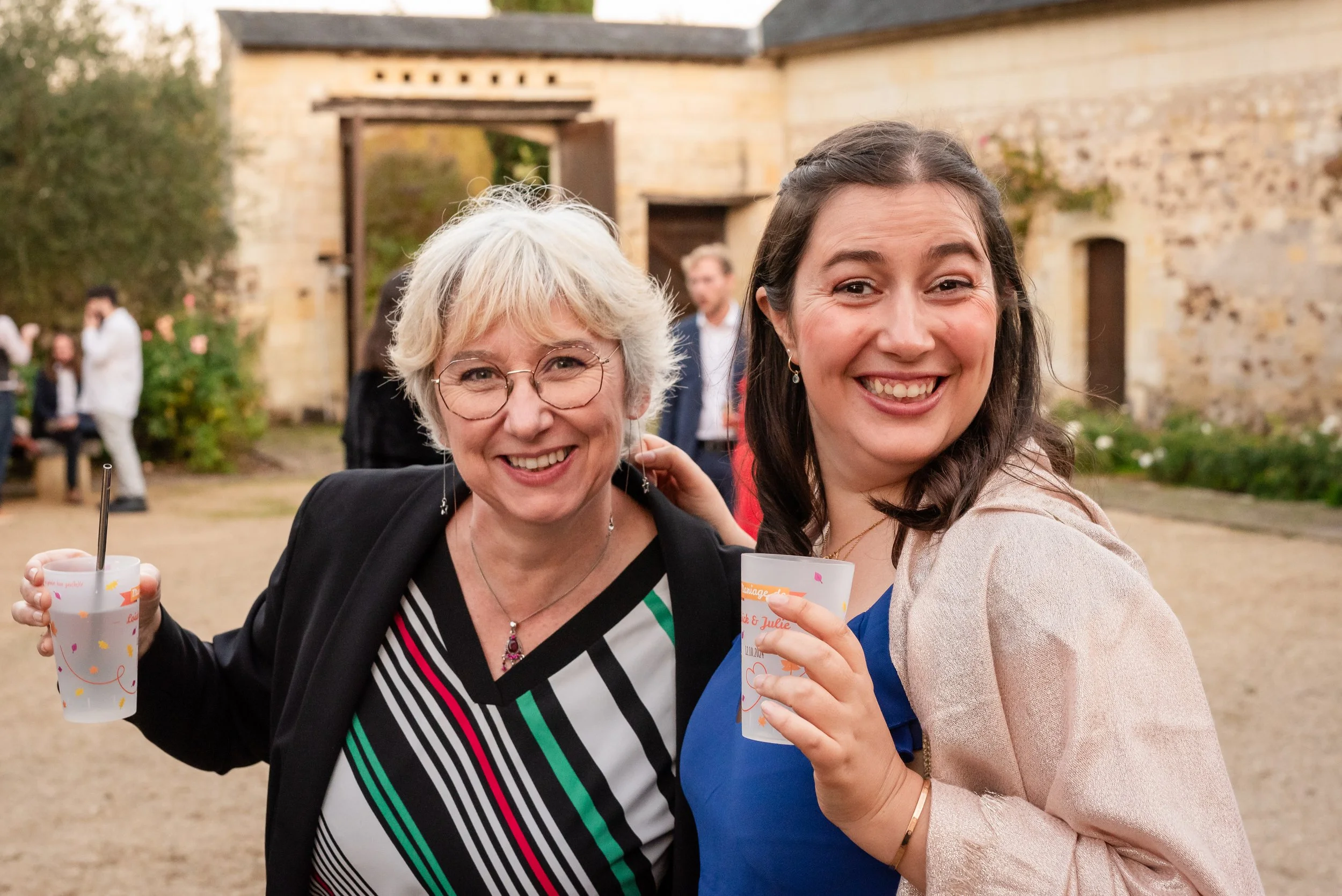 Deux femmes souriantes en extérieur, tenant des gobelets, lors d'une célébration ou fête, avec un bâtiment en pierre en arrière-plan.