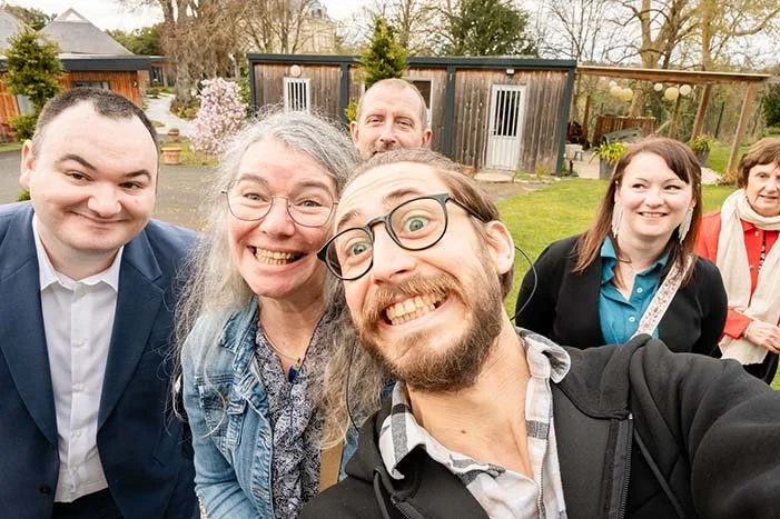Groupe de personnes souriantes posant pour une selfie en extérieur près d'un bâtiment en bois, au printemps.