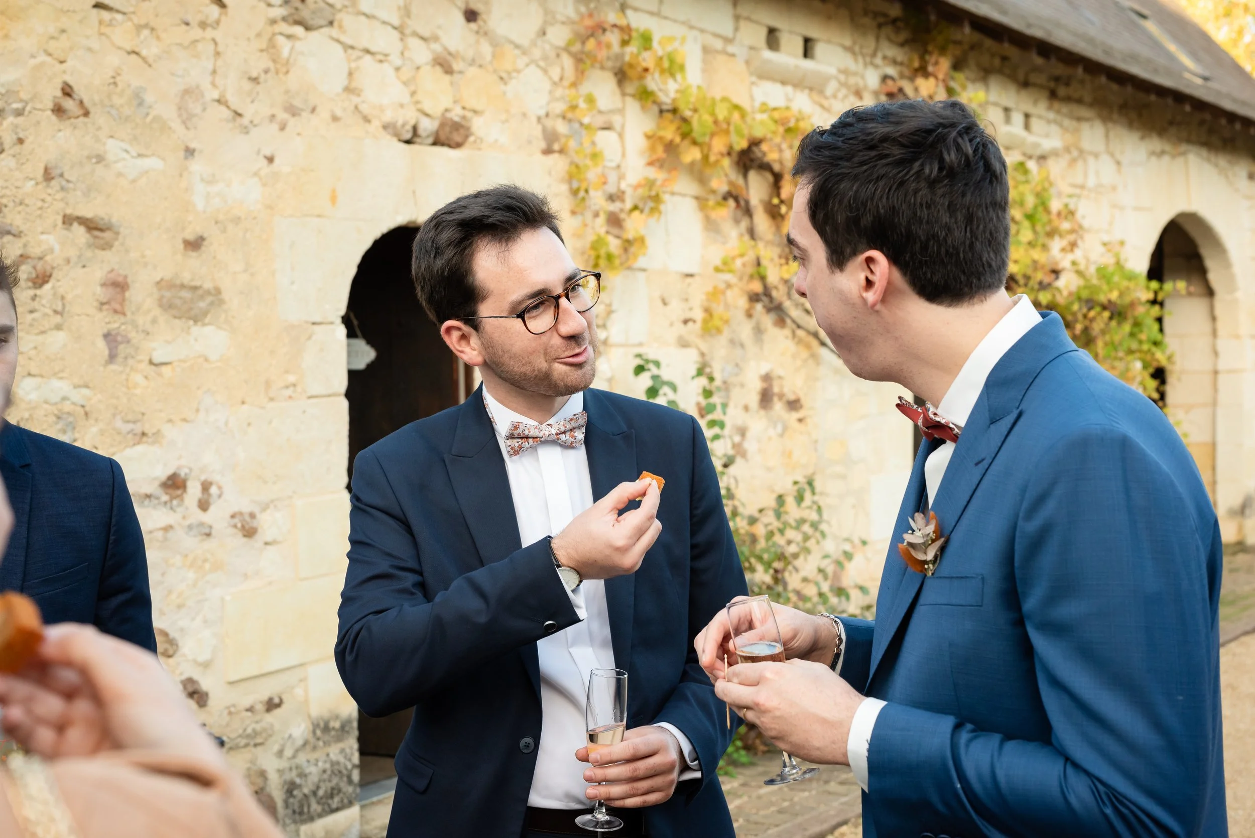 Deux hommes en costume discutent lors d'une réception en extérieur, tenant des verres de champagne, dans un décor de bâtiment en pierre avec des vignes.